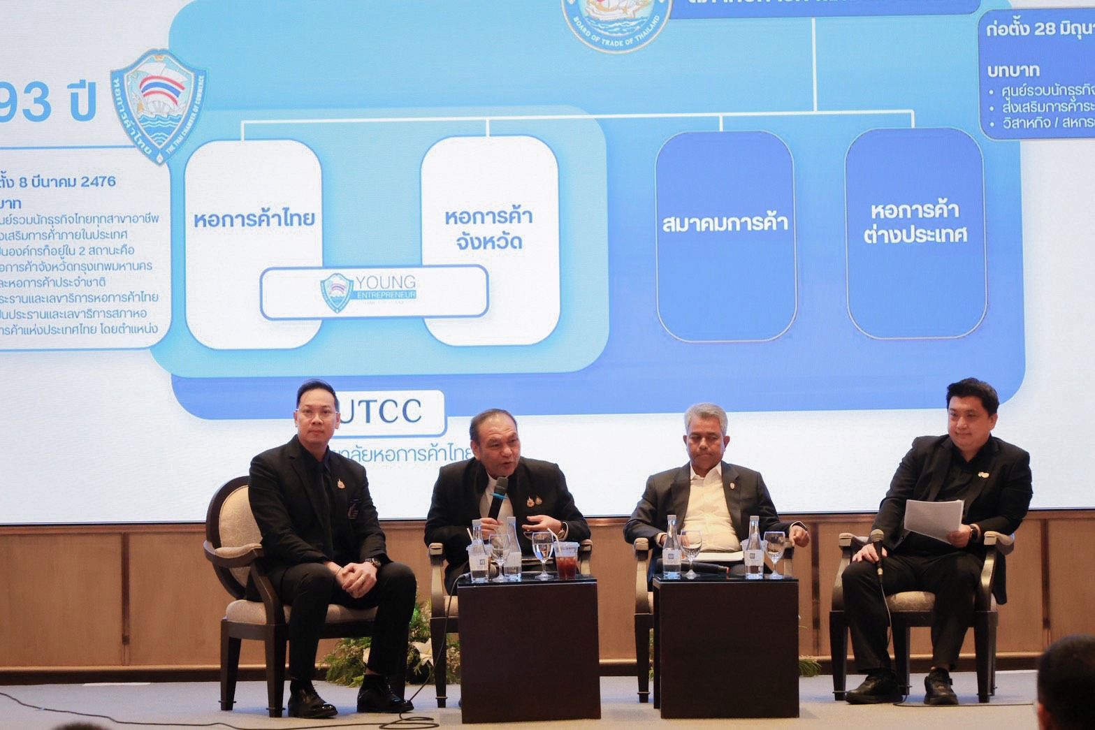 Four men conduct a panel discussion with a large screen displaying an organizational diagram.