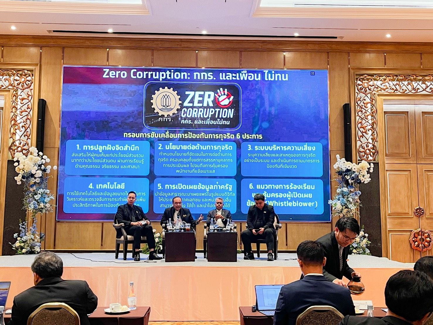 Panelists discuss anti-corruption policies in a conference room with a large screen displaying Thai text.