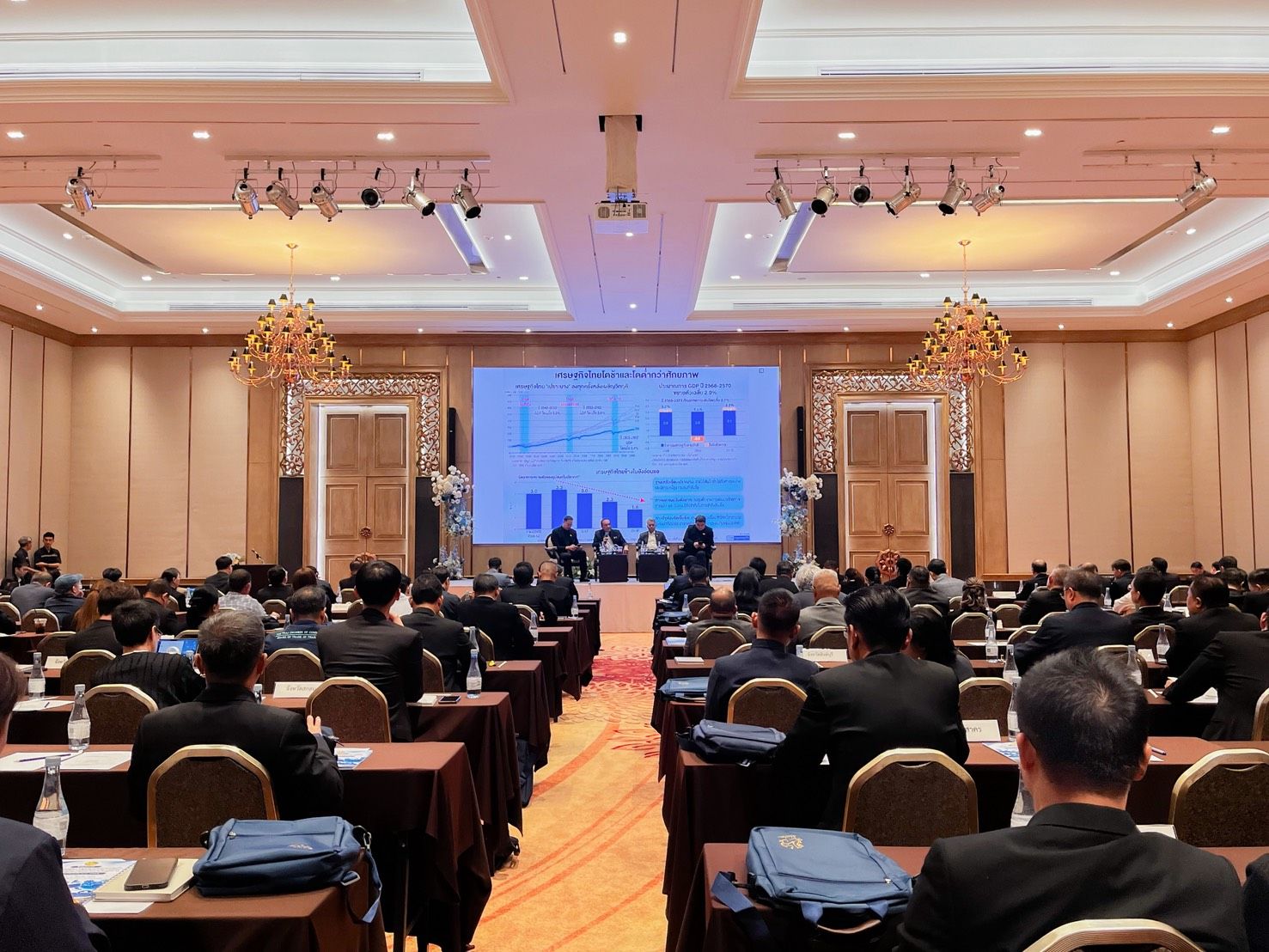 Attendees at a large conference in a ballroom, facing a stage with a presentation screen.