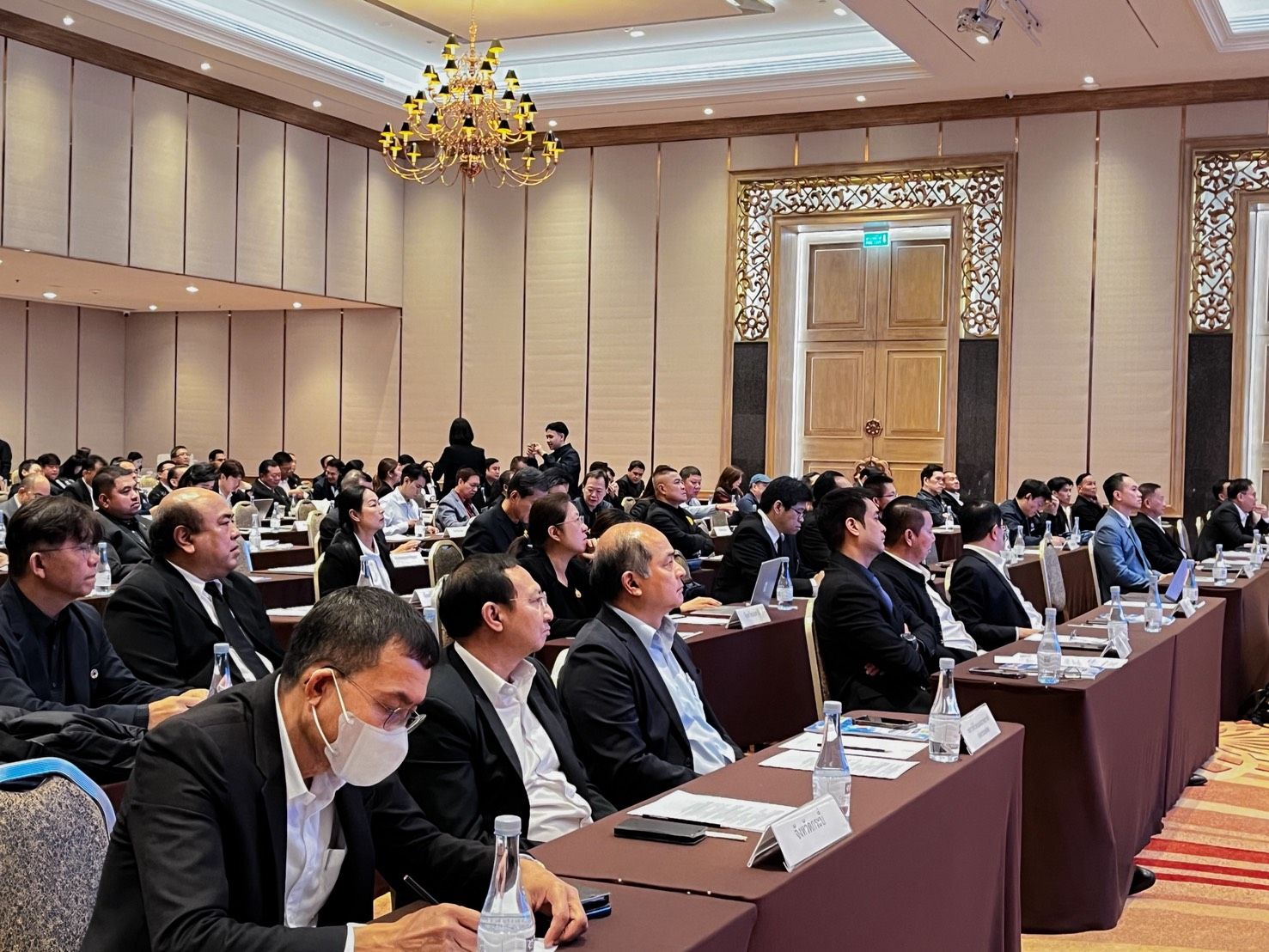 A large conference room filled with attendees seated at long tables, watching a presentation.