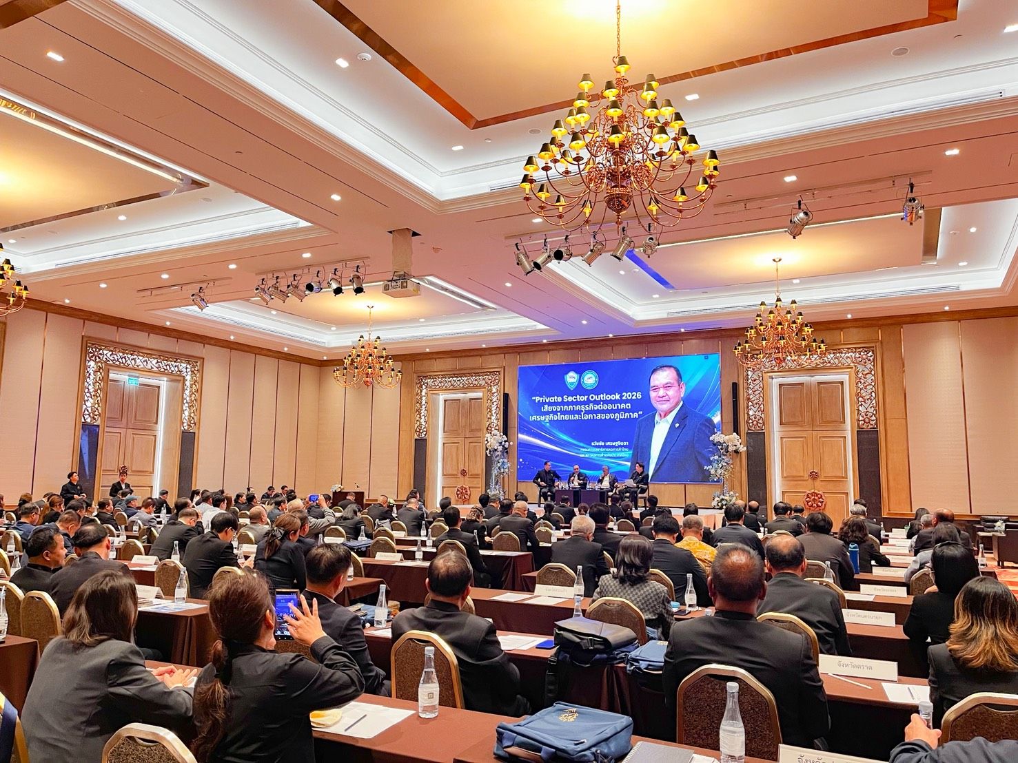 Audience in a large conference hall watching a speaker on a big screen.