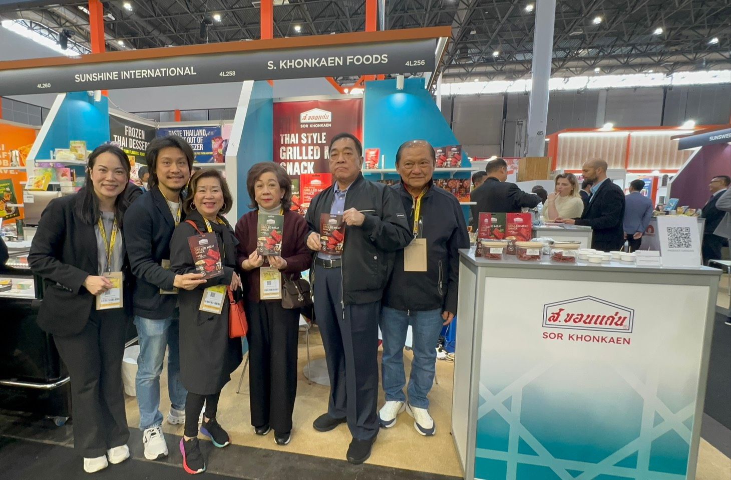 Seven smiling people hold food products at a Nagoya brand booth in a trade show.