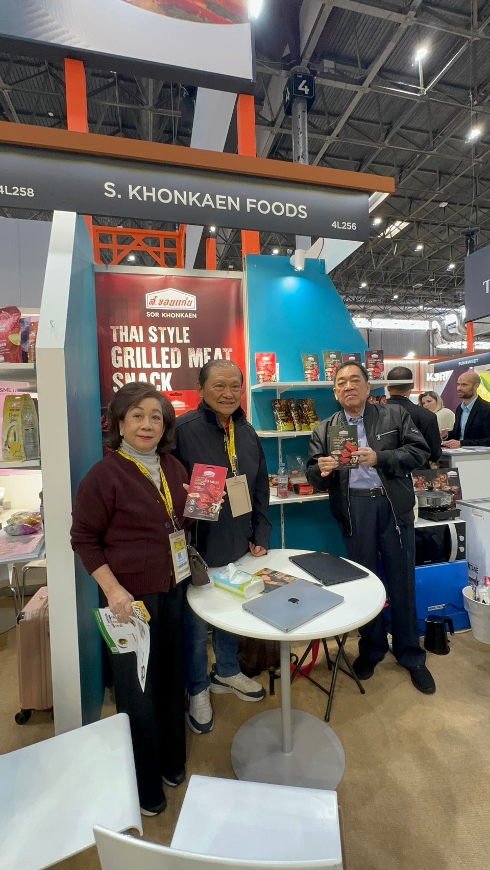 Three people stand at a trade show booth with a sign reading 'OUR LITTLE CHICKEN HELPS'.