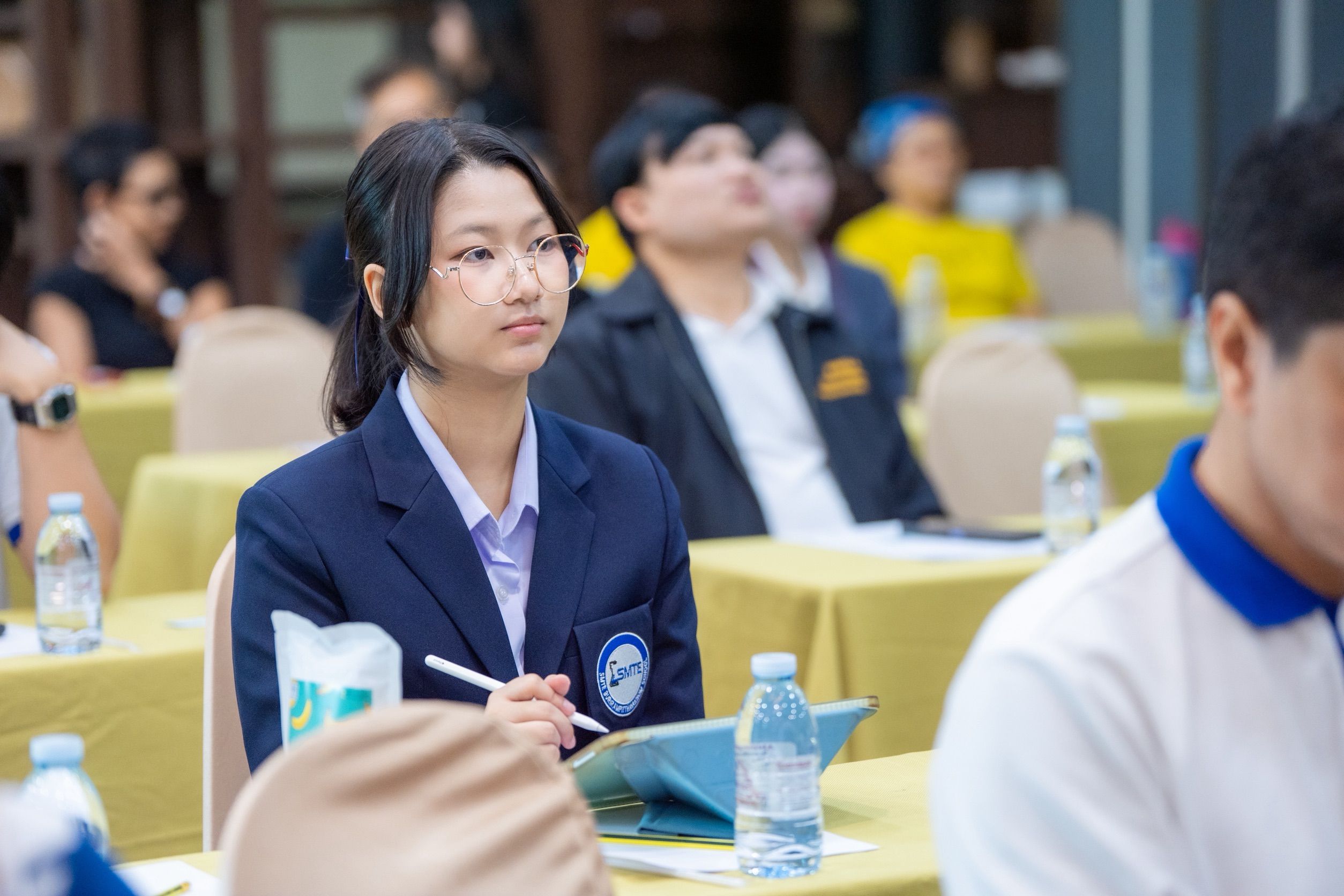 Young Asian woman in a blue blazer attentively listens during a conference session.
