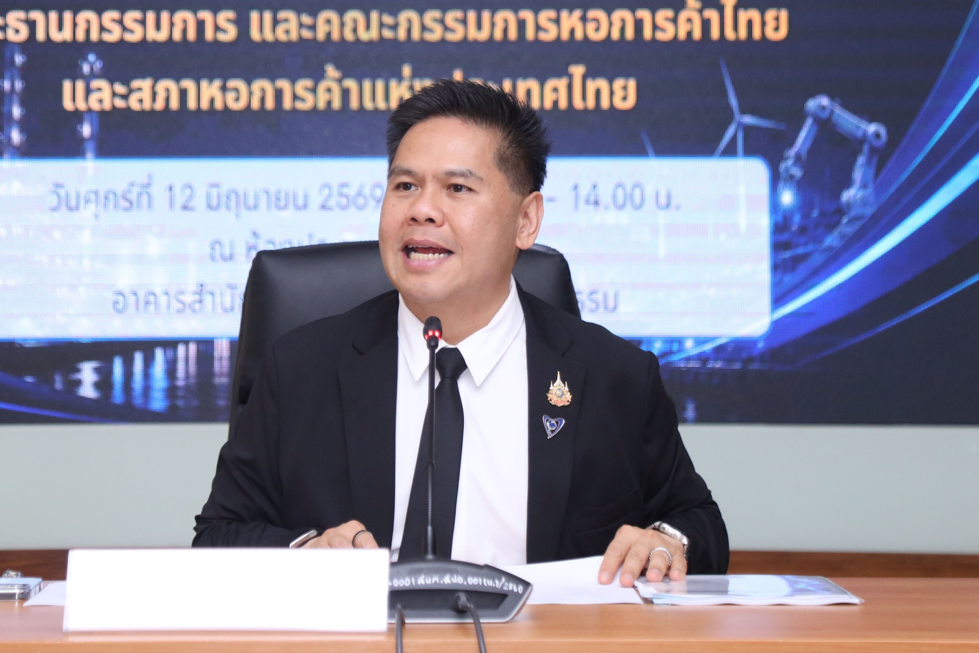 Man in dark suit speaking at a formal event with Thai text on a screen.
