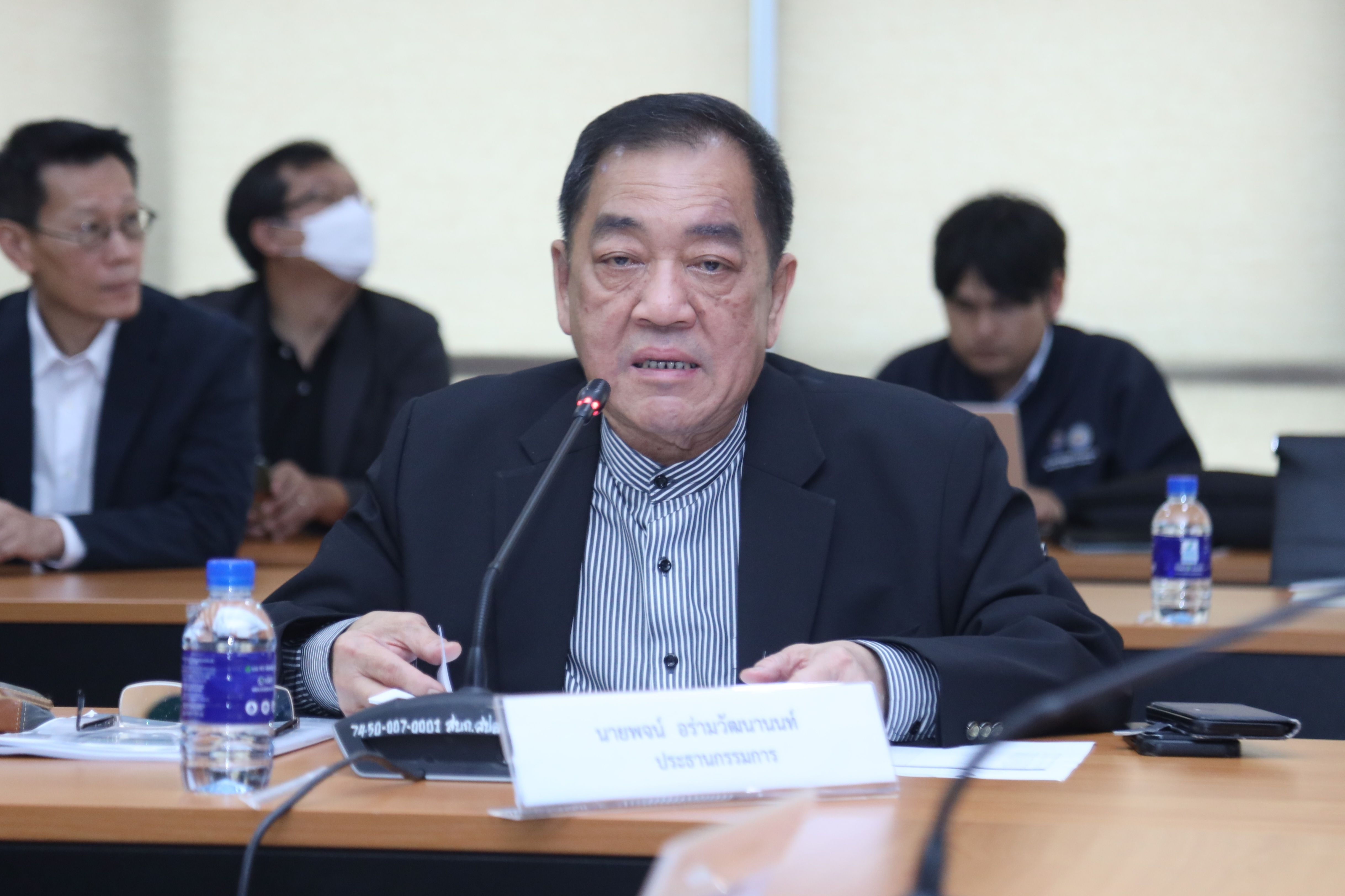 Older Asian man in a dark suit jacket speaks into a microphone at a conference table.