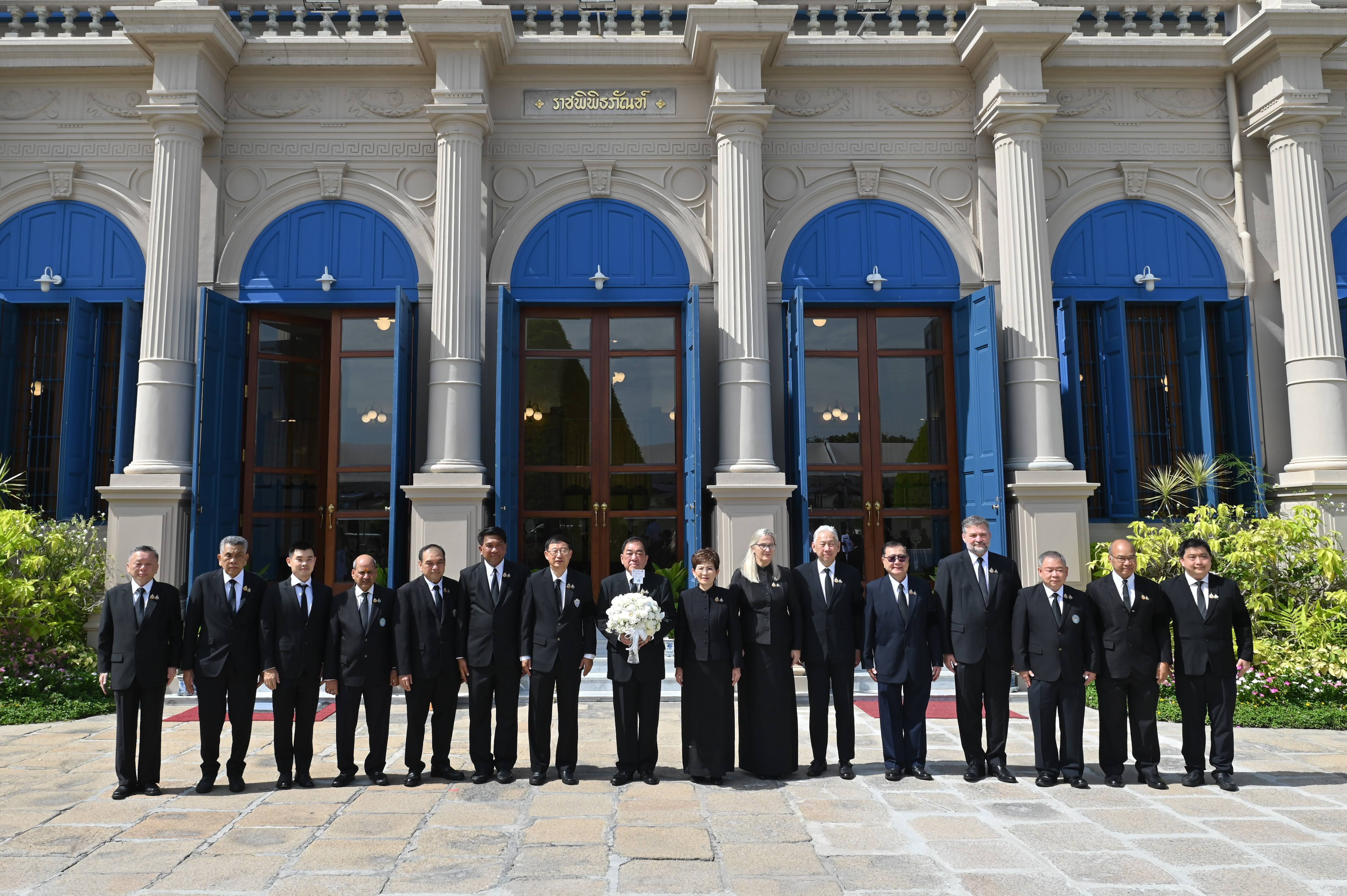 A formal group of 14 people, some holding white flowers, stand before a grand building.