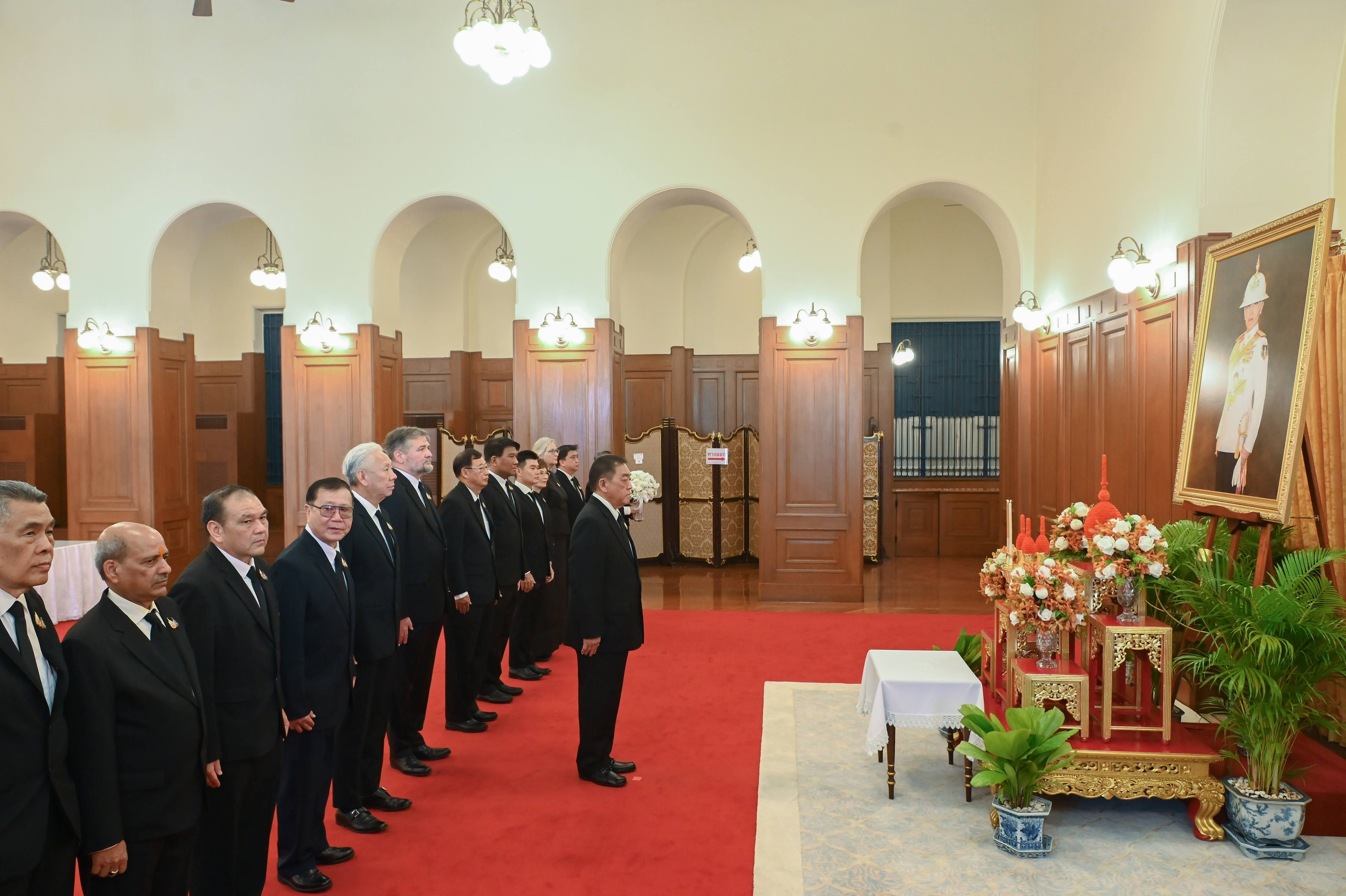 Men in suits stand formally in a room, facing a royal portrait and ceremonial display.