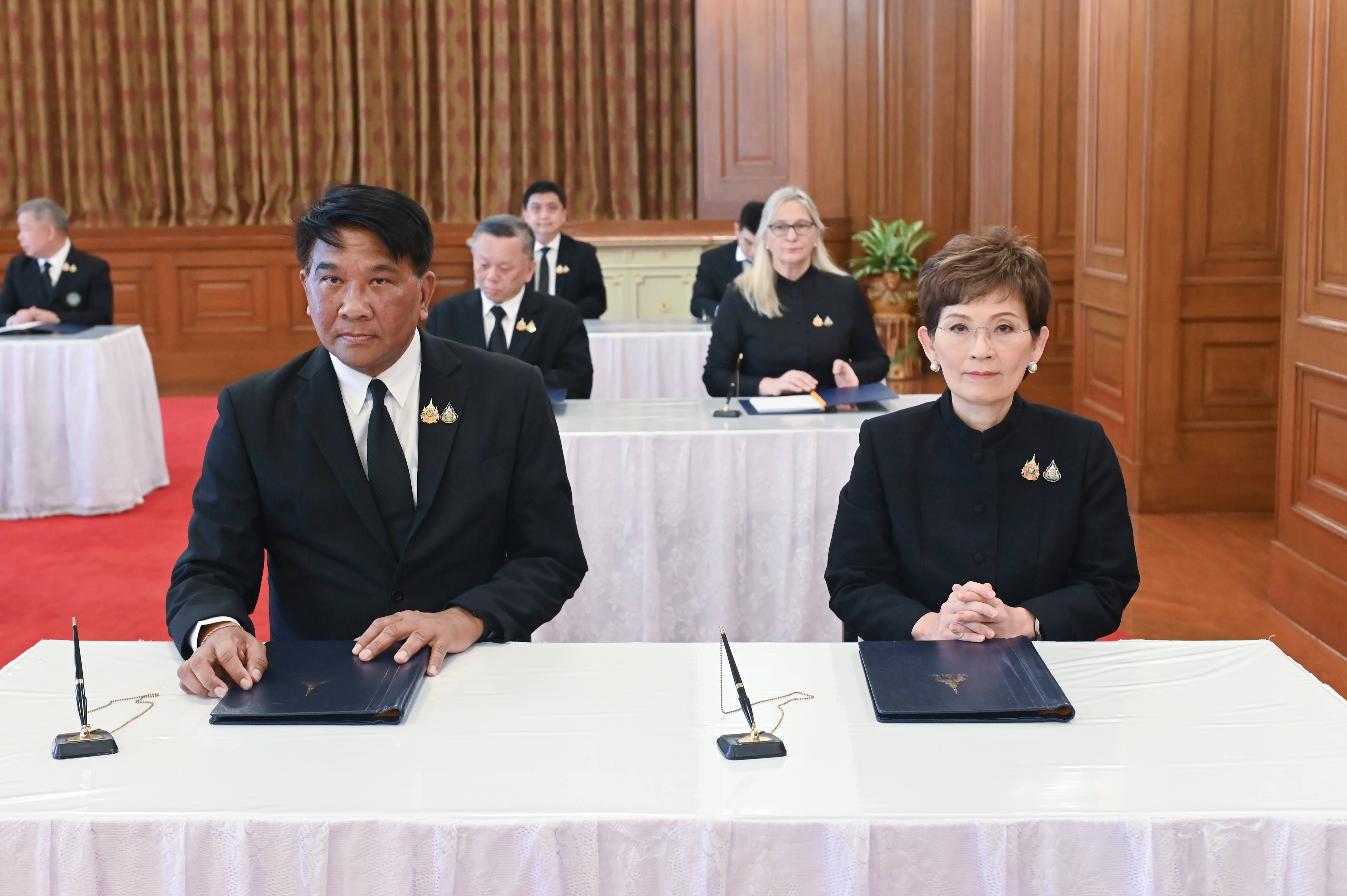 Two officials in black formal attire seated at a white table with folders.