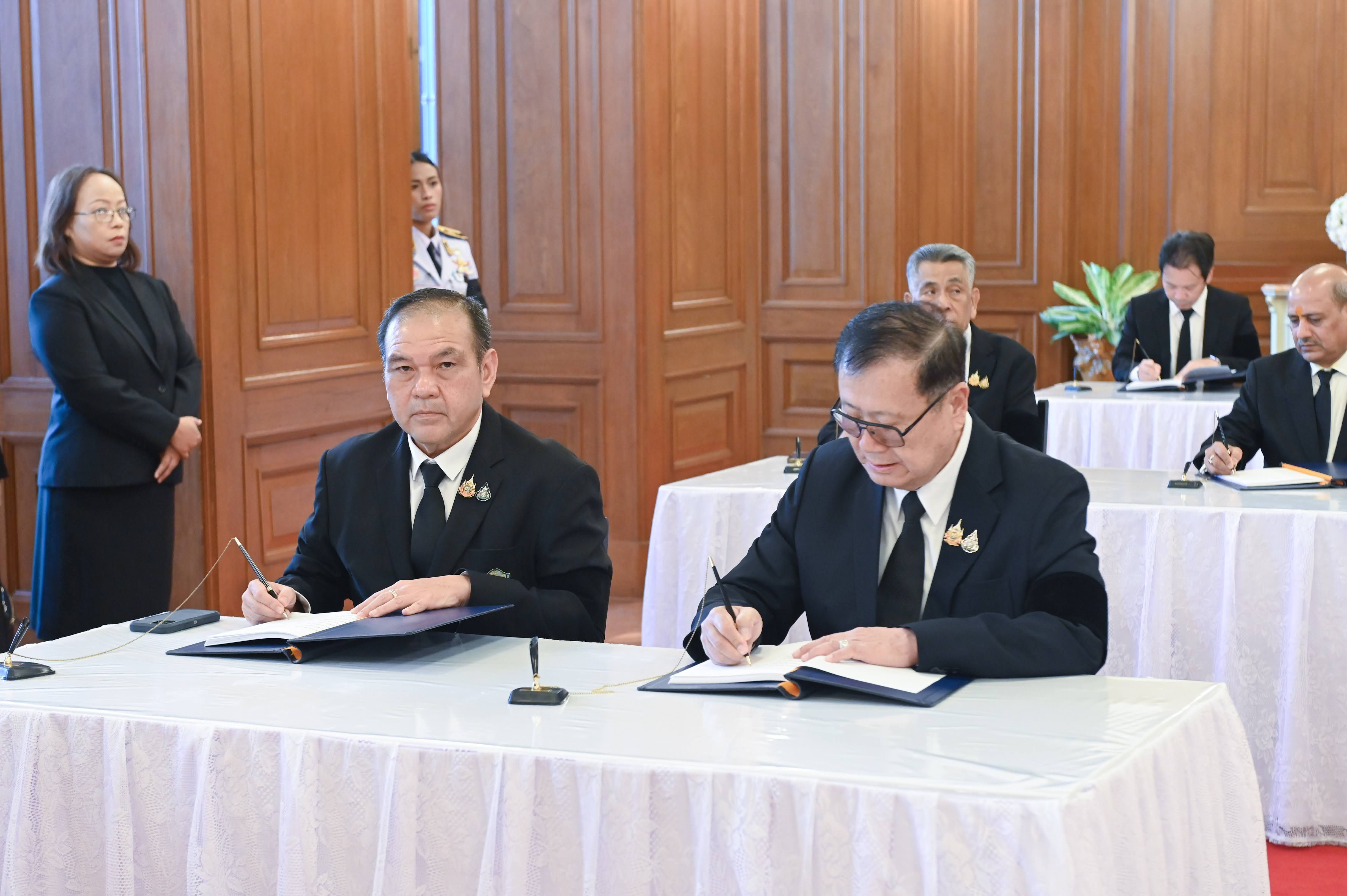Two men in dark suits formally sign documents at a long white table.