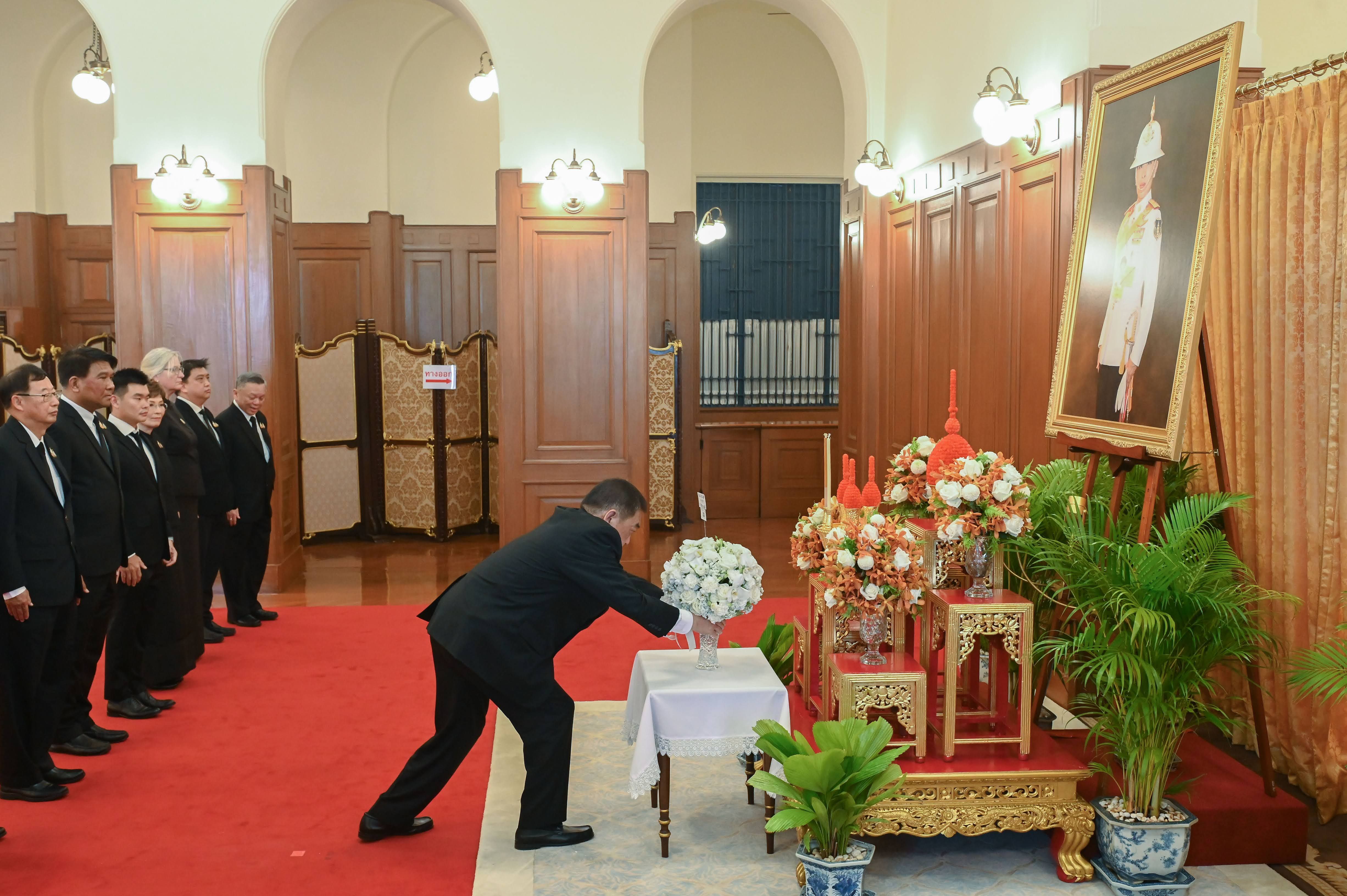 Man in suit places white flowers on a table during a formal ceremony with other attendees.