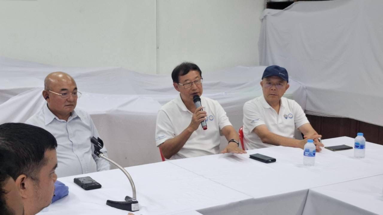 Three men at a meeting, one speaking into a microphone at a white table.