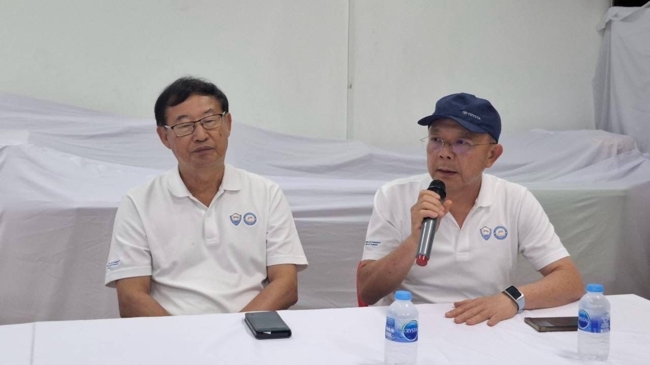 Two men in white polo shirts, one speaking into a microphone at a table.