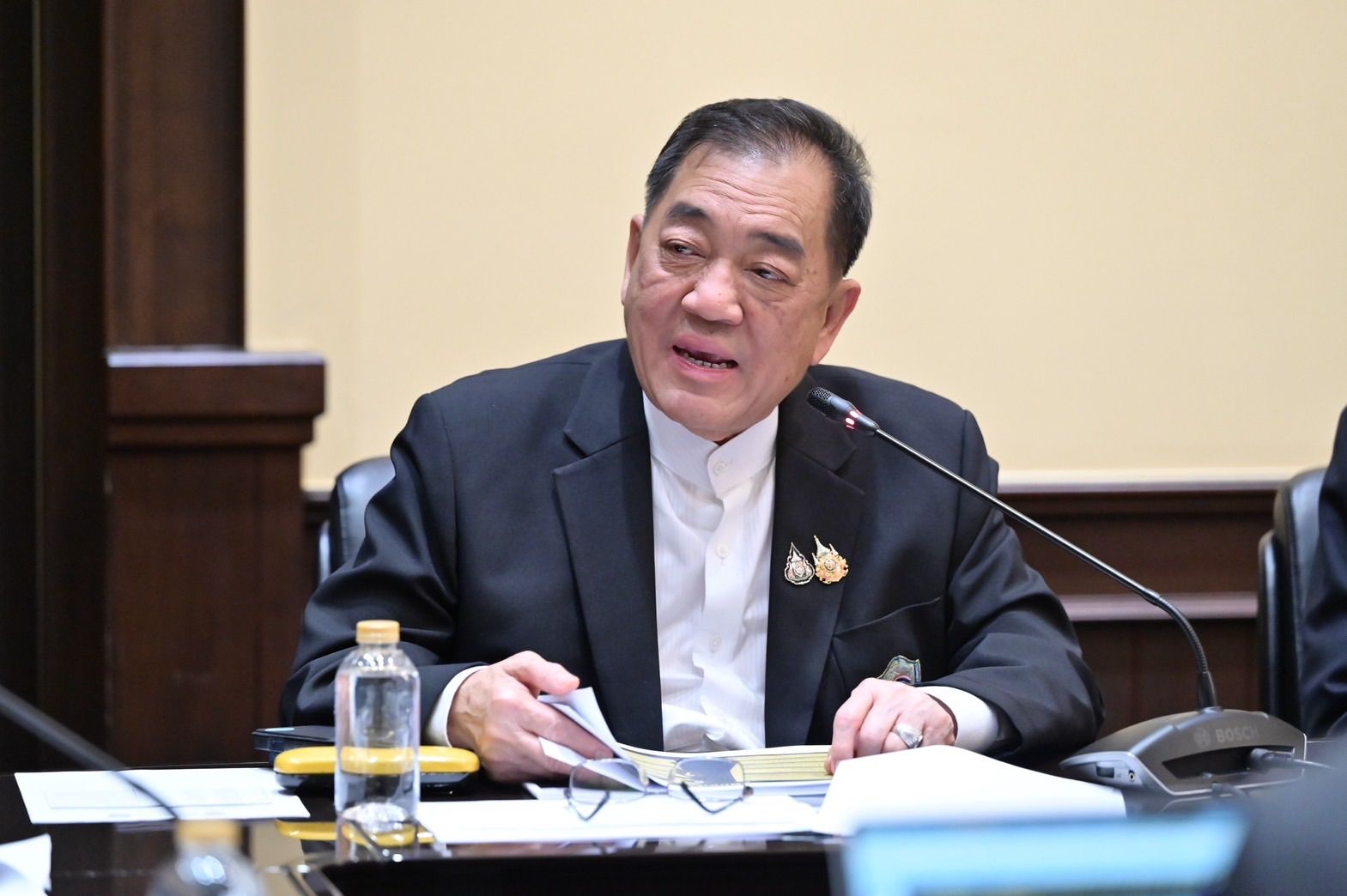 An Asian man in a black suit speaks at a conference table, holding papers.