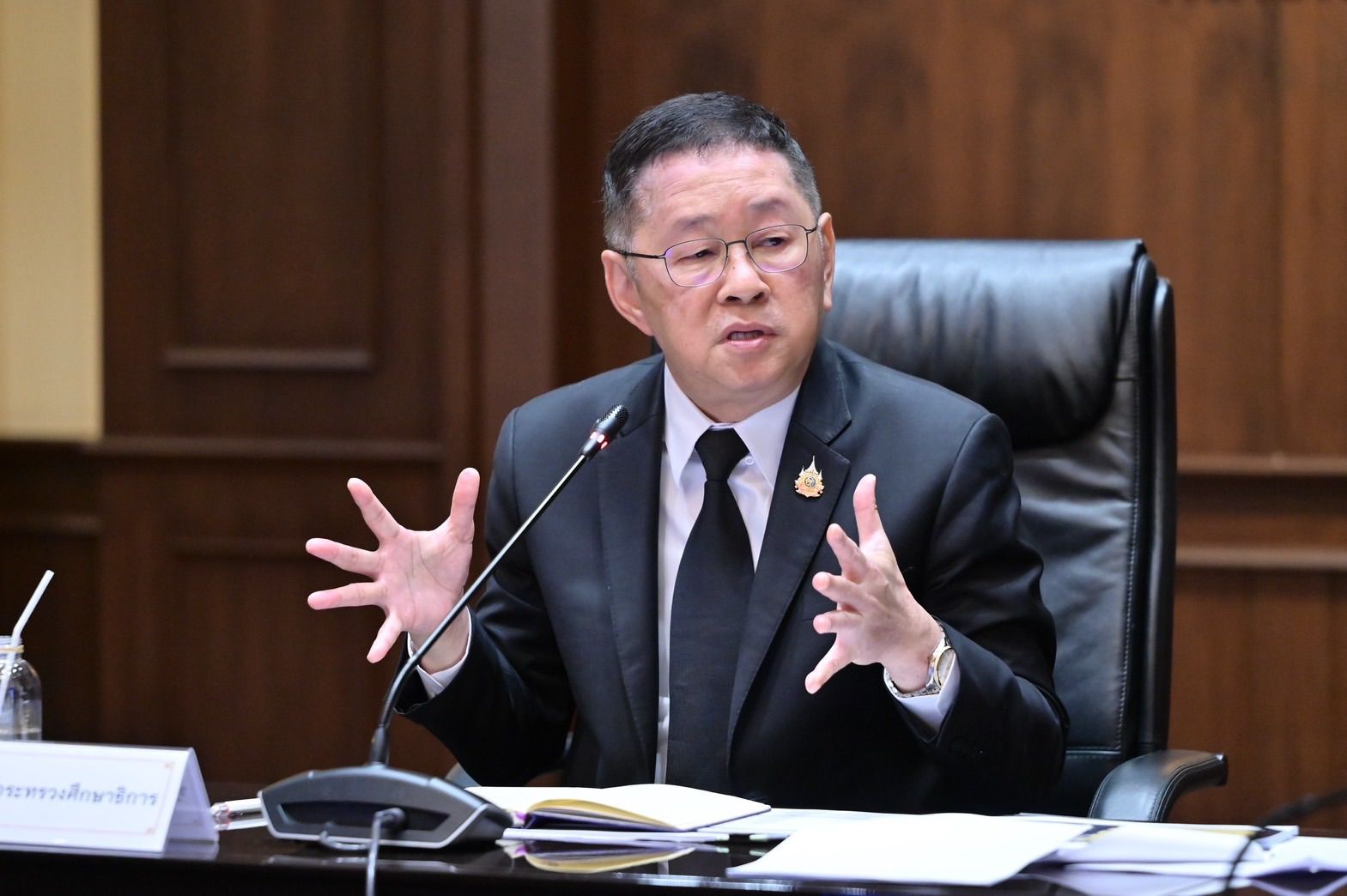 Man in dark suit and glasses speaks animatedly, gesturing with both hands at a conference table.