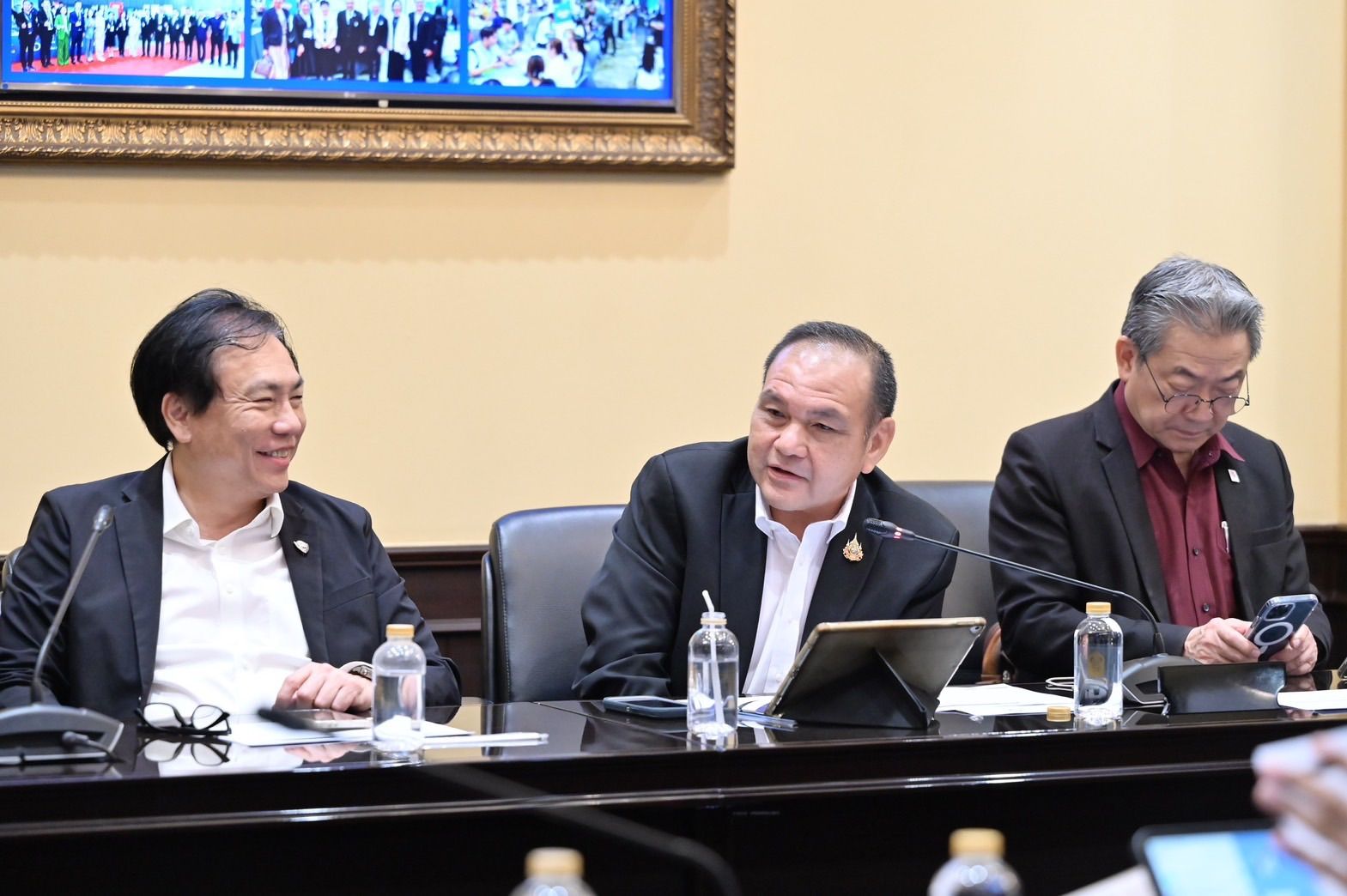 Three men in suits at a meeting table; one smiles, one speaks, one uses a phone.