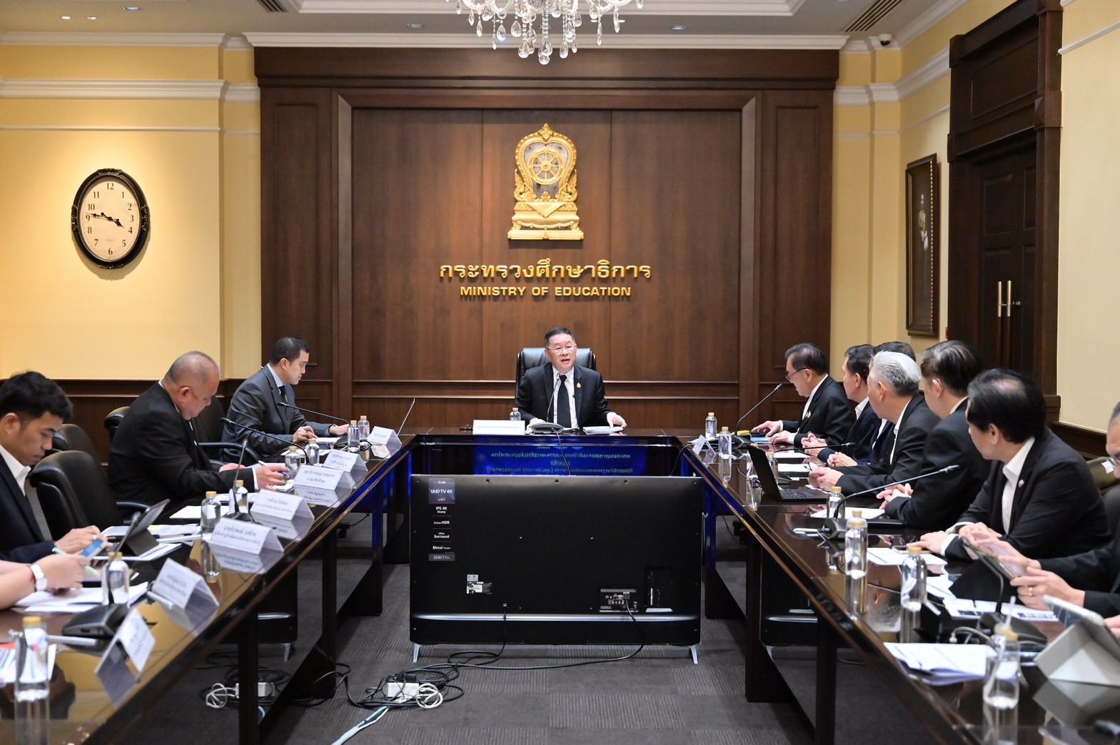 Officials hold a formal meeting in a wood-paneled room with a long table and golden emblem.