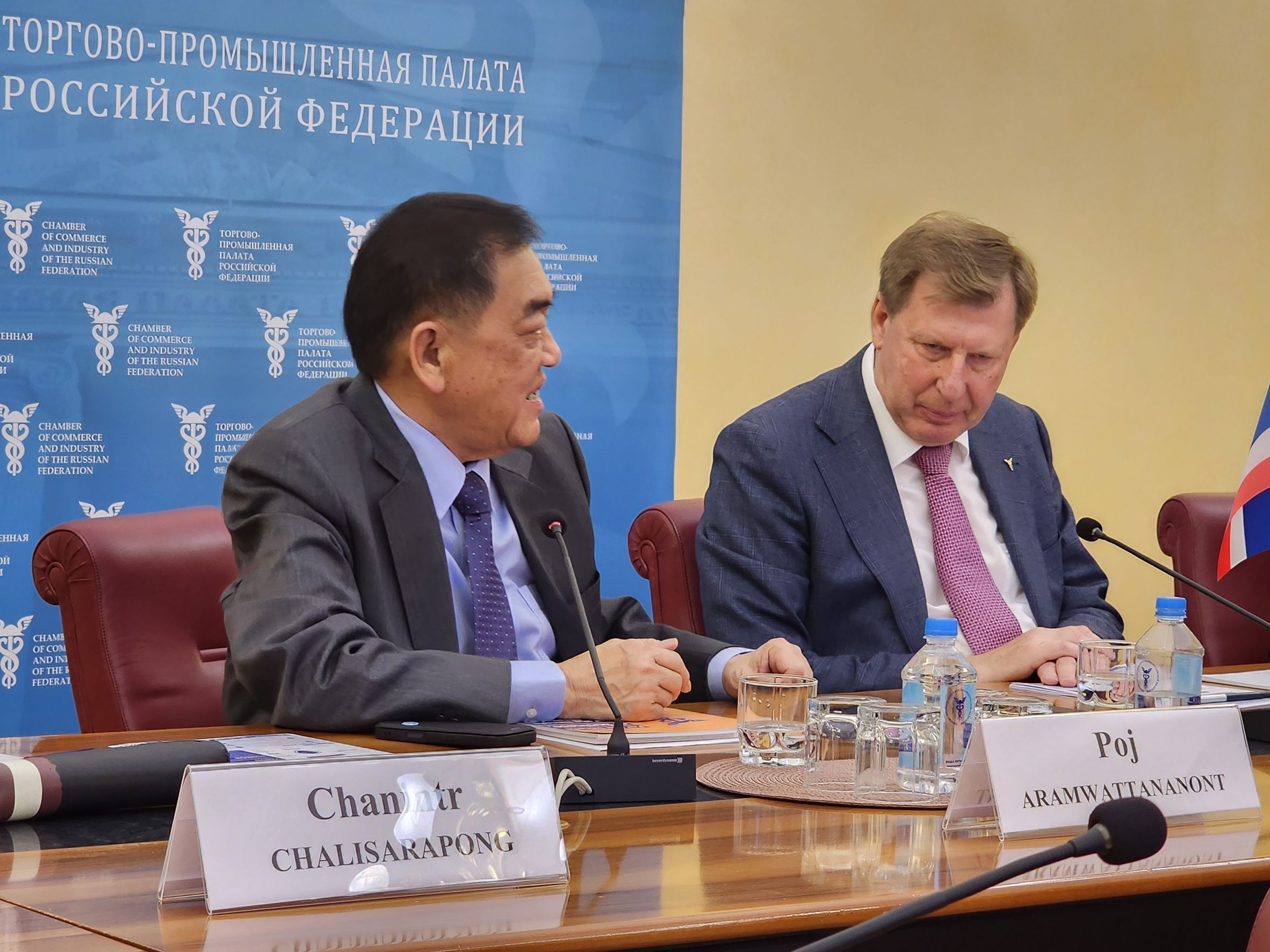Two men in suits attend a formal meeting, one smiling and looking at the other.