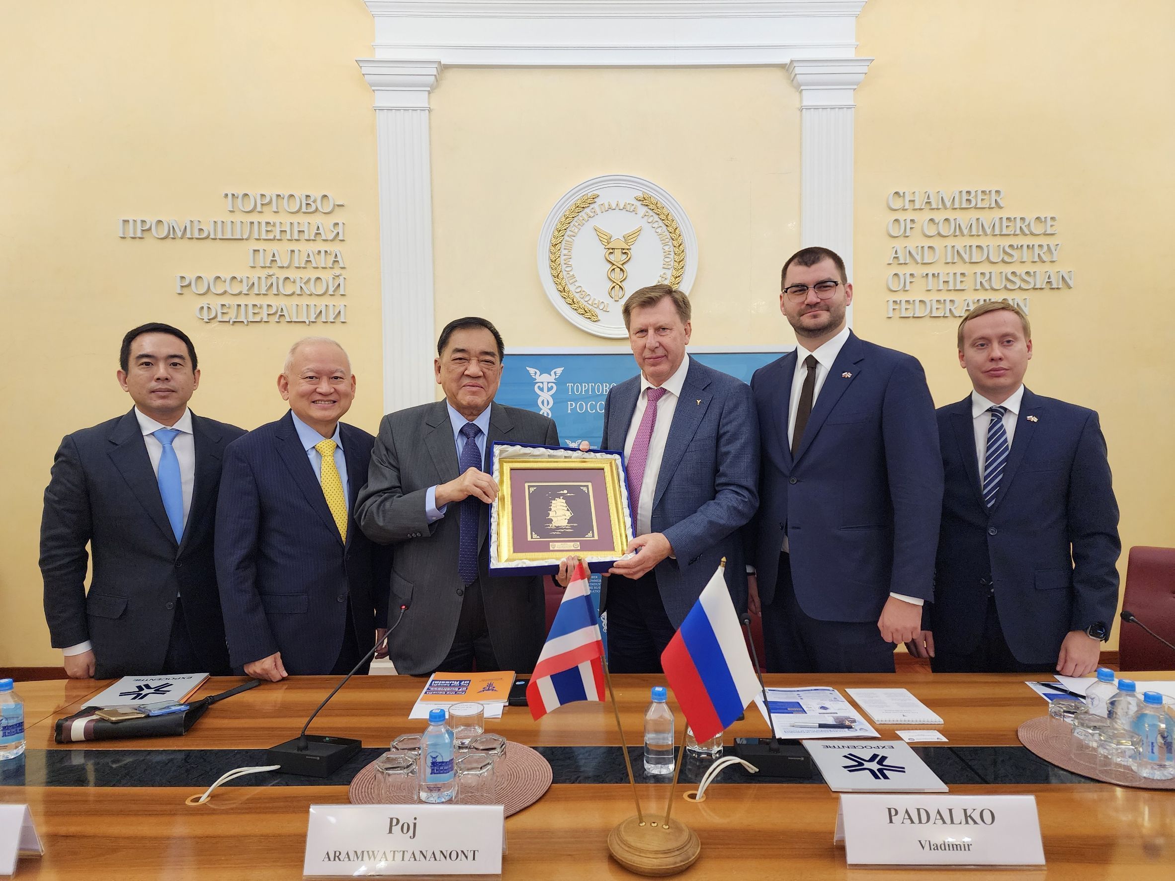 Six men in suits exchange a framed gift during an international meeting with Thai and Russian flags.