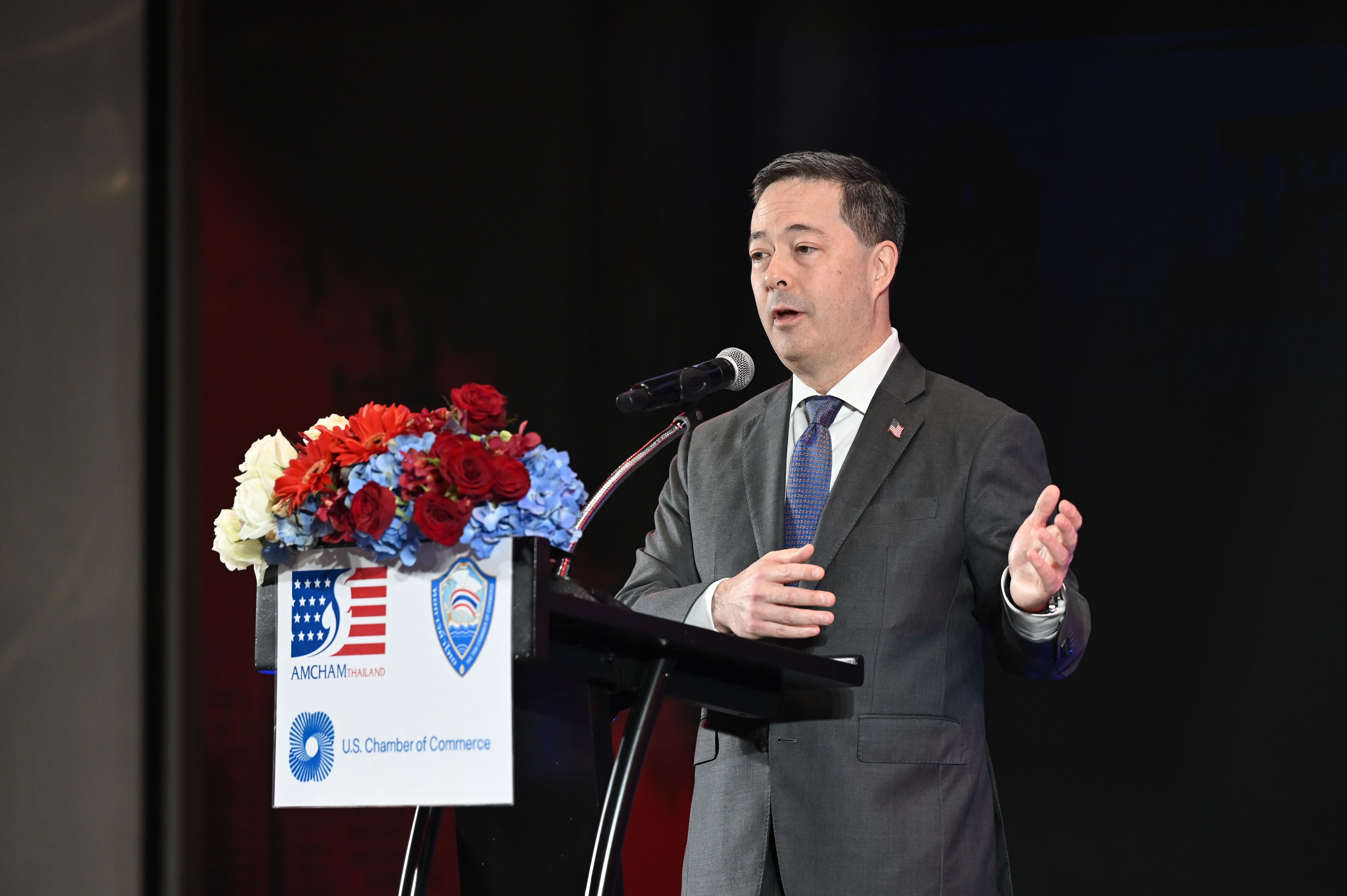 Man in suit speaking at a podium with 'U.S. Chamber of Commerce' sign and floral arrangement.
