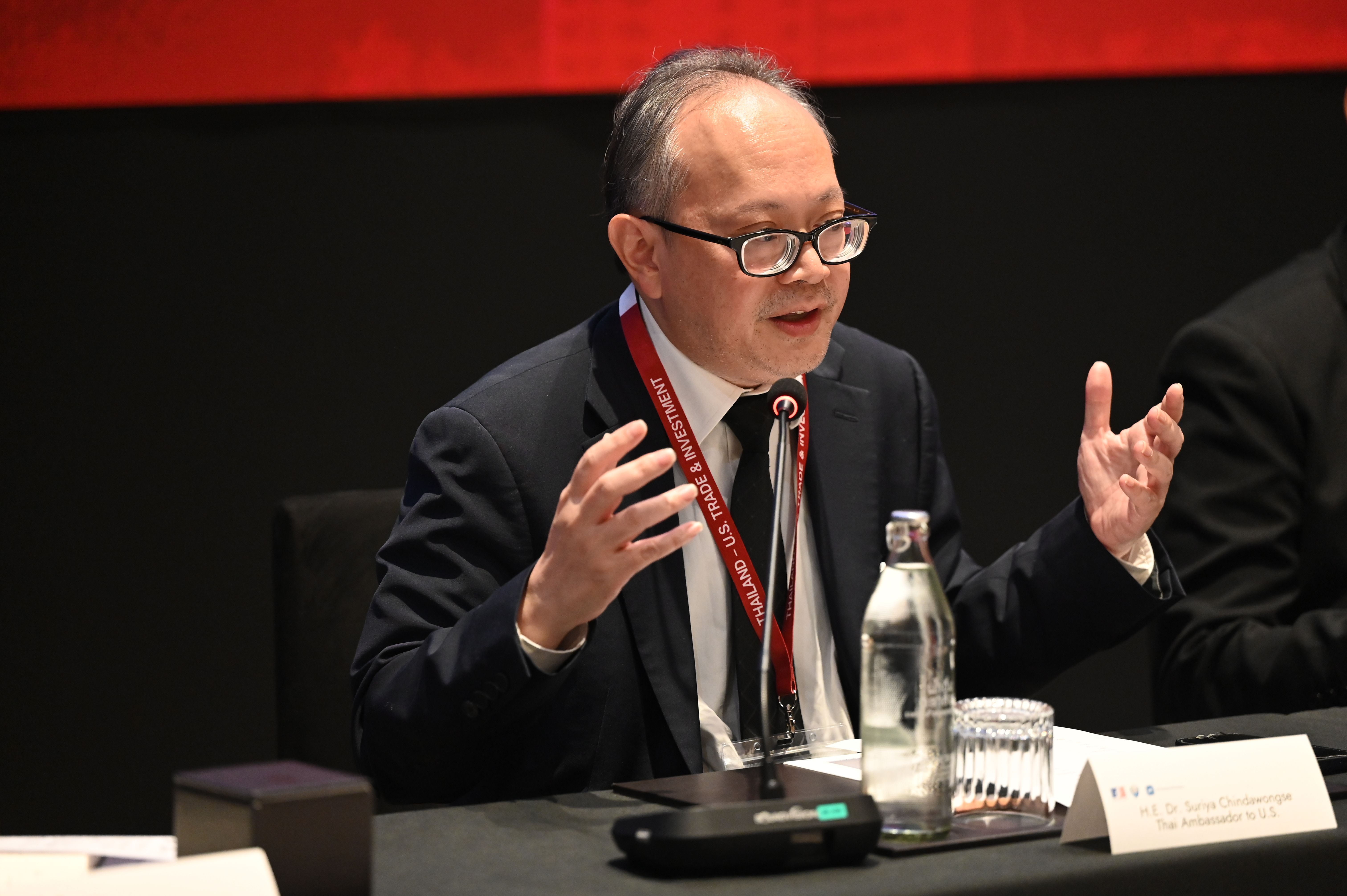Man in glasses and suit speaking passionately into a microphone at a conference table.