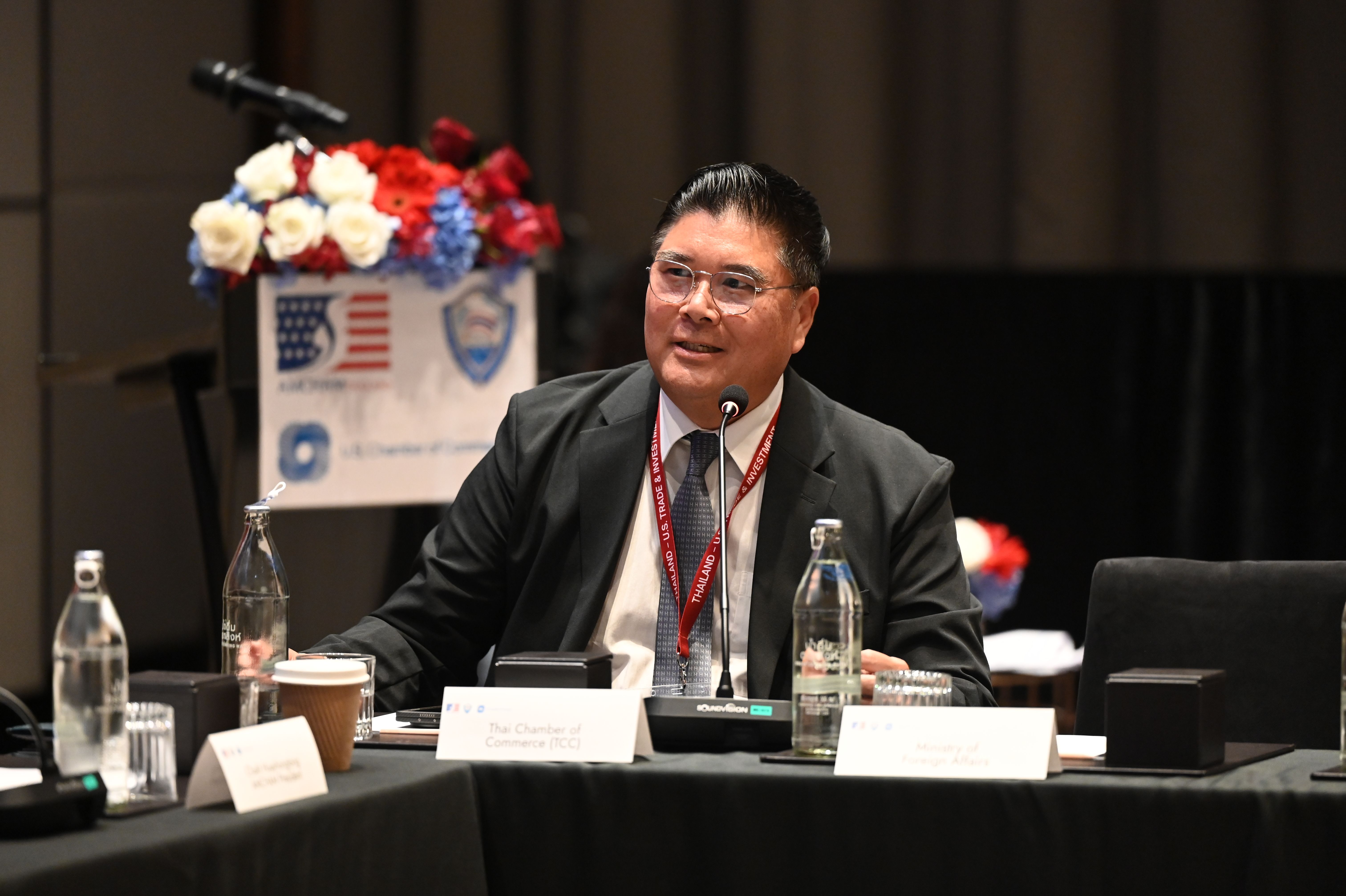 Asian man in a dark suit and glasses speaking at a conference table with a microphone.