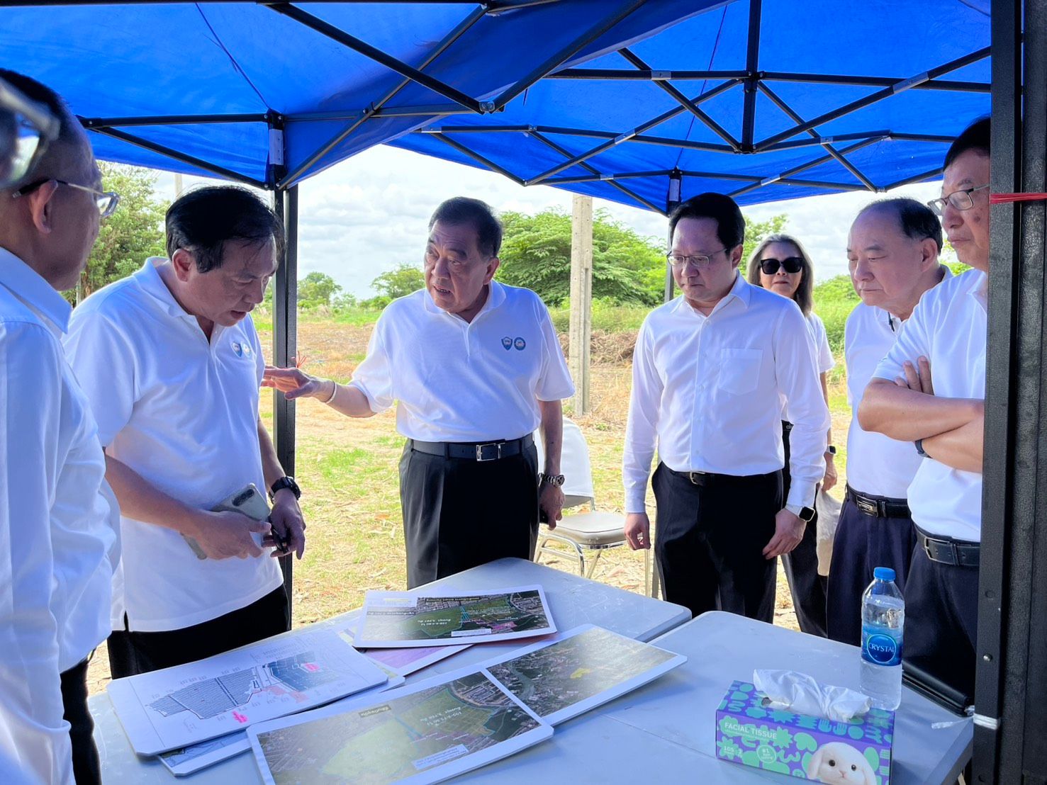 Officials in white shirts discuss documents under a blue canopy tent outdoors.