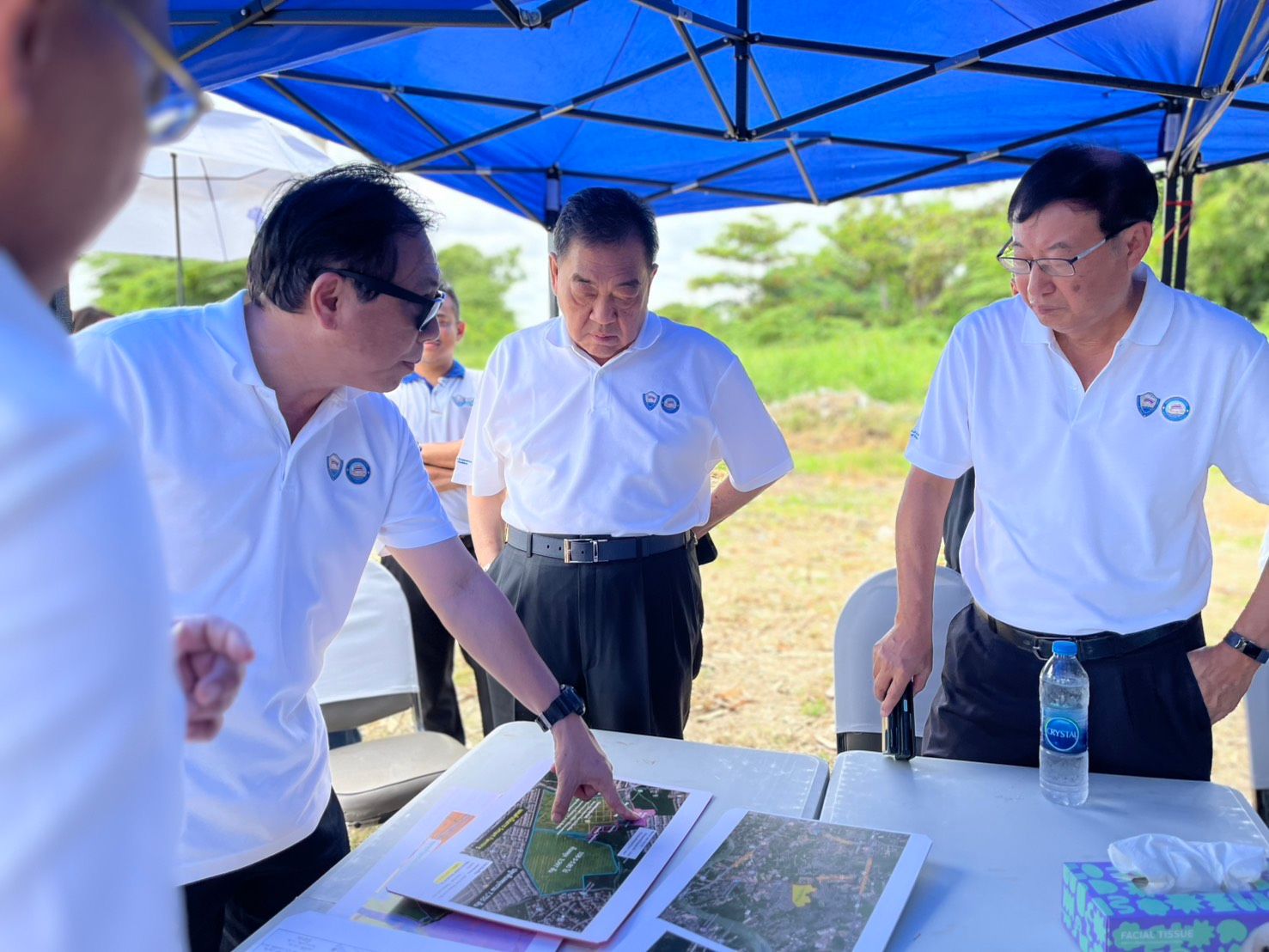 Three men in white shirts review documents under a blue canopy outdoors.