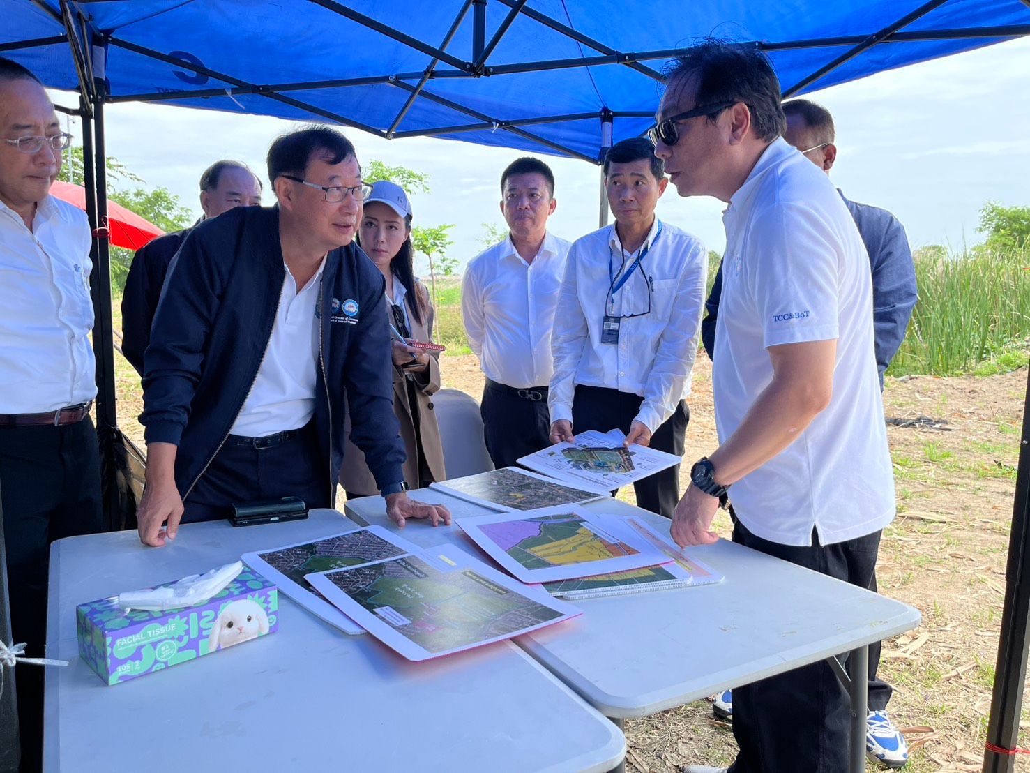 Two men discuss documents on tables under a blue tent with others observing outdoors.