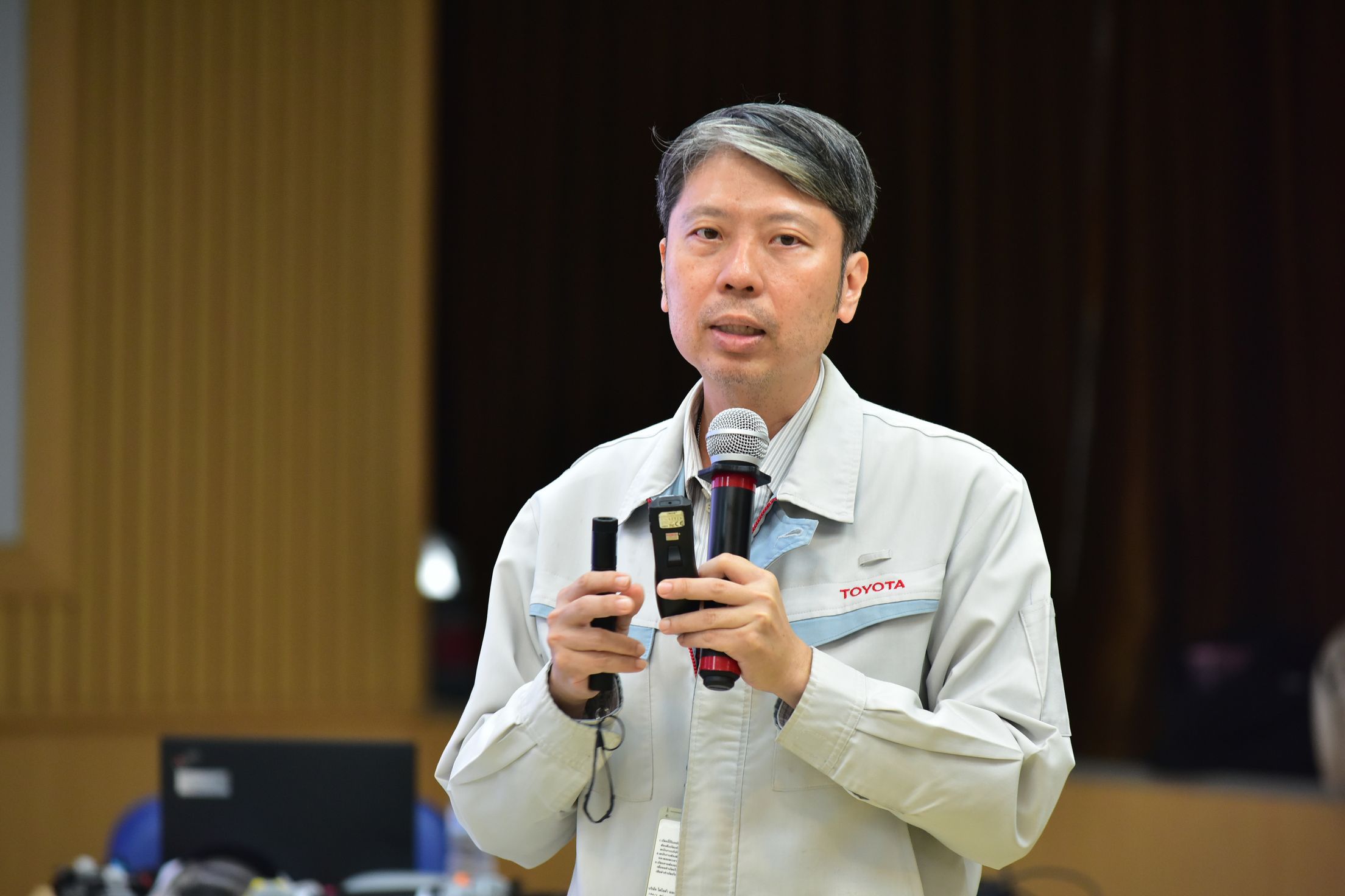 Man with grey hair speaking into a microphone, holding a device during a presentation.