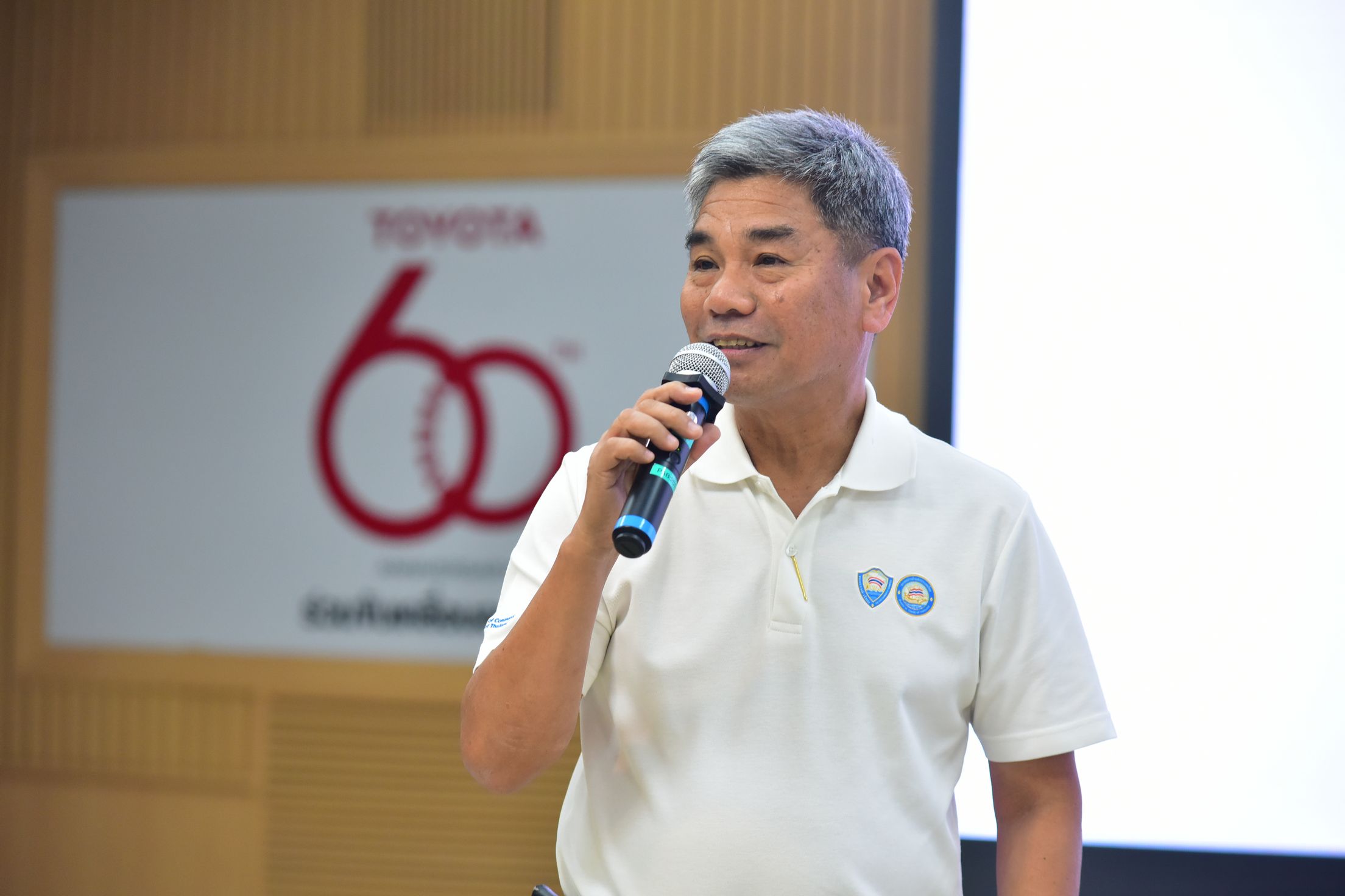 Man speaking into a microphone with a Toyota 60th anniversary logo in the background.