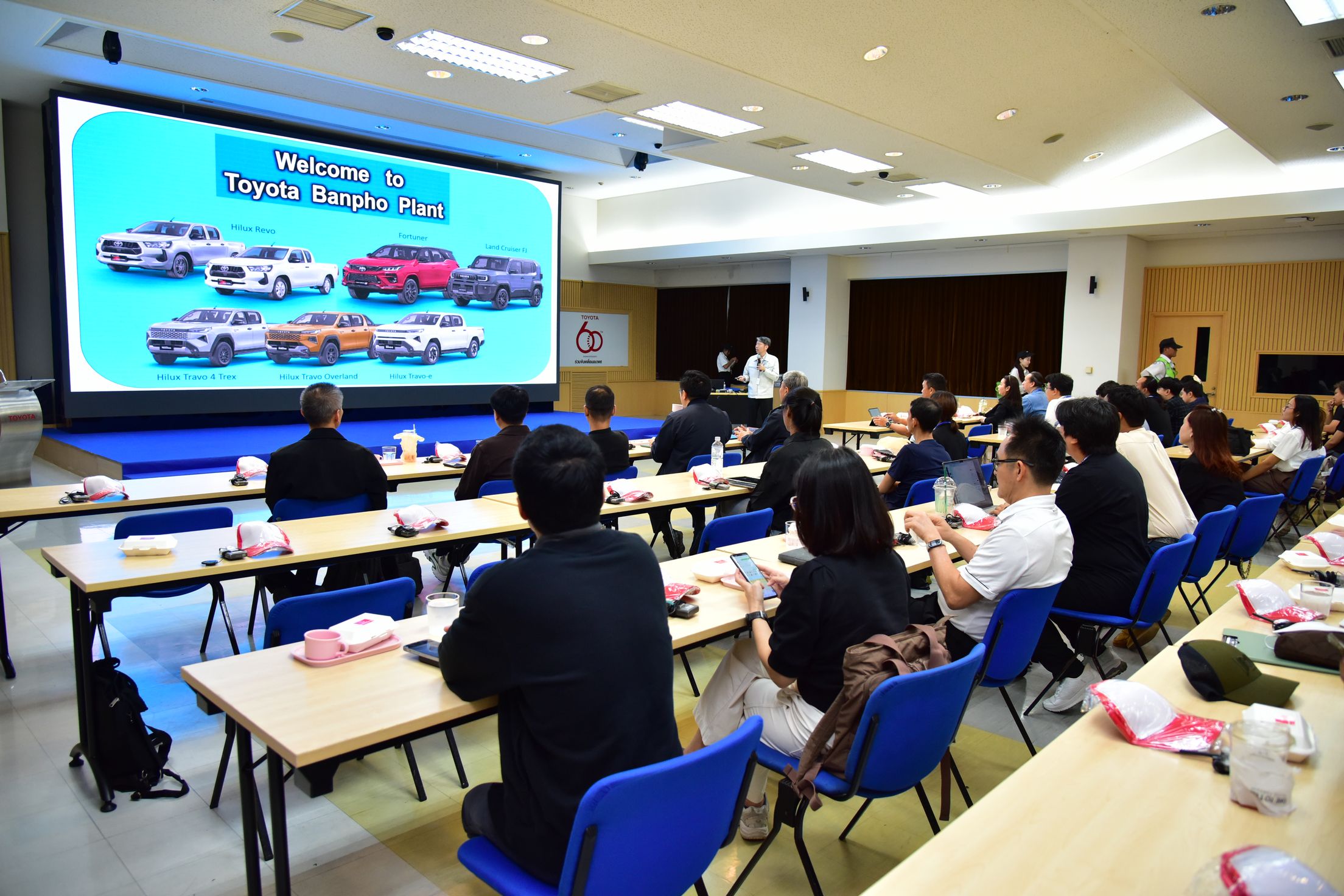 Attendees watch a large screen presentation displaying multiple car models in a workshop setting.