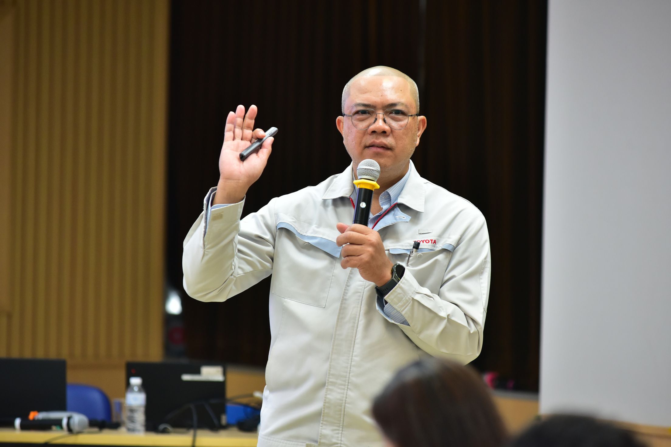 A bald man wearing glasses speaks into a microphone while raising his hand during a presentation.