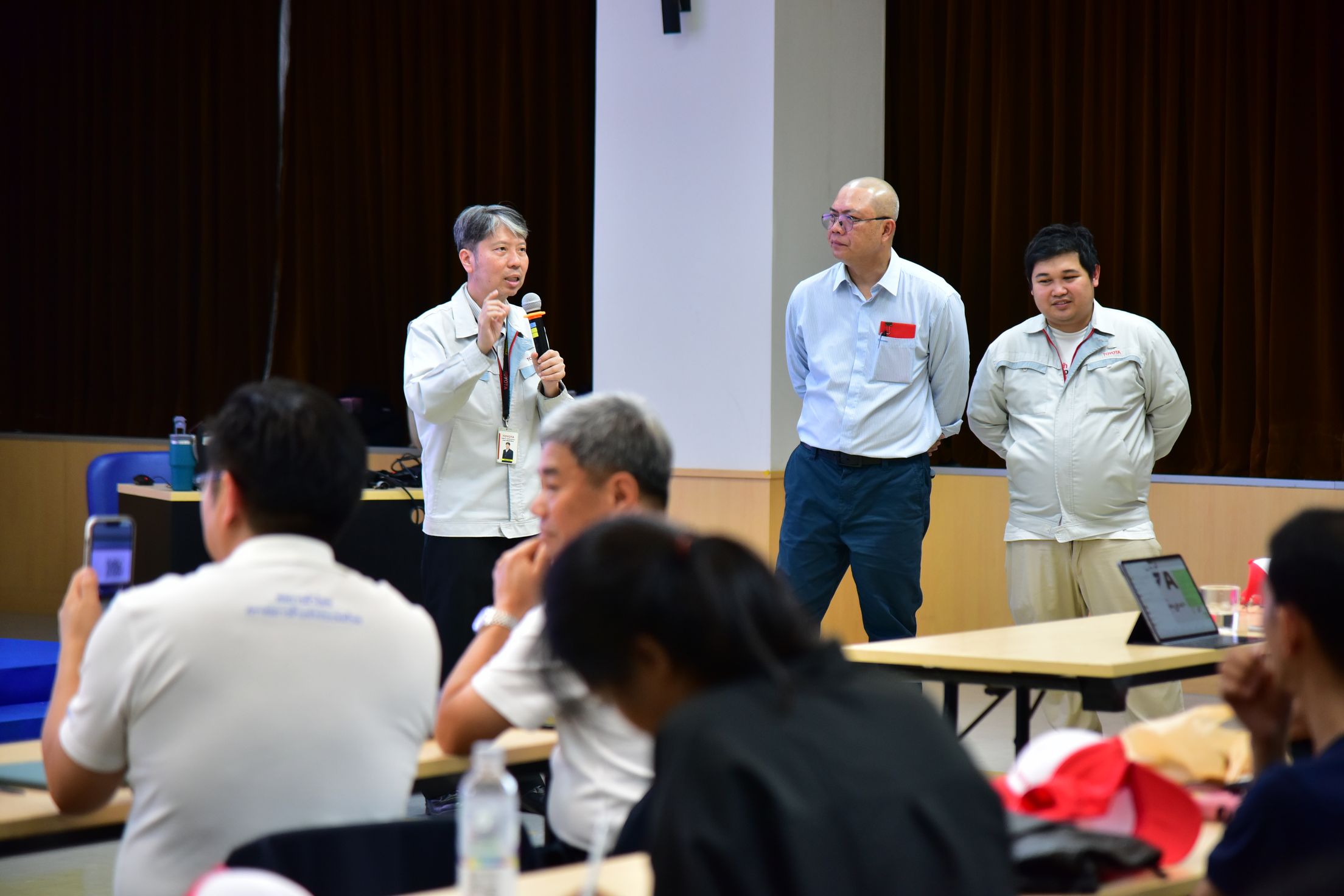 Speaker addresses an audience at a conference, with two men observing from behind.