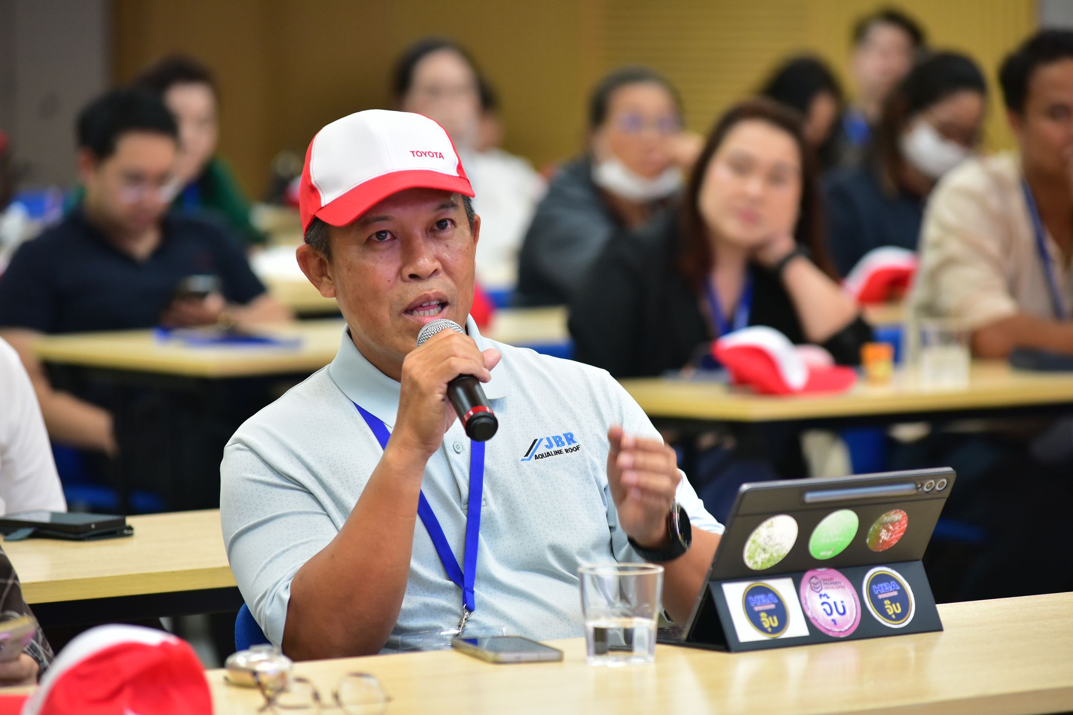 Man in red and white cap speaks into microphone during a conference.