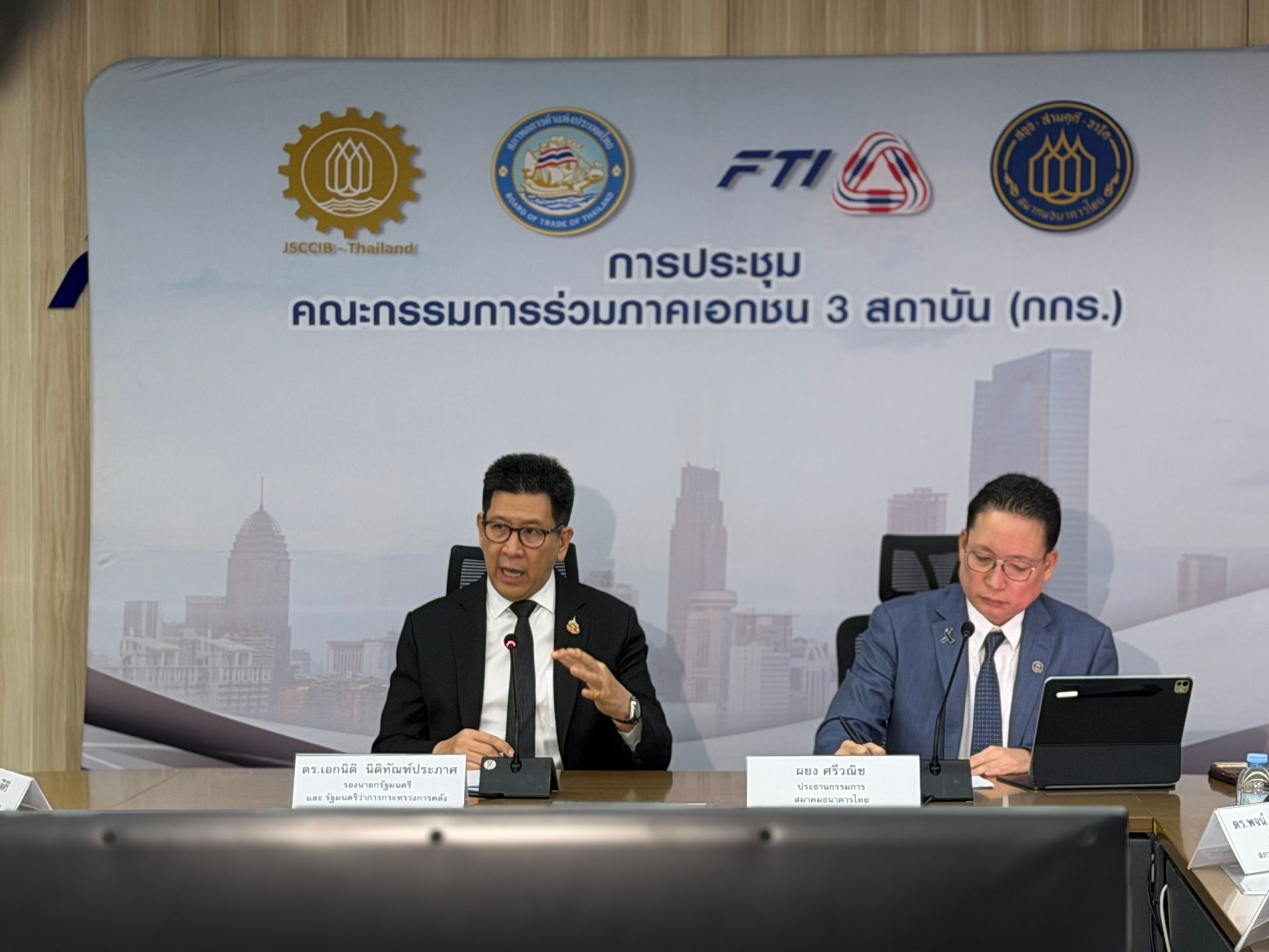 Two men in suits at a conference table, one speaking with hand gestures.