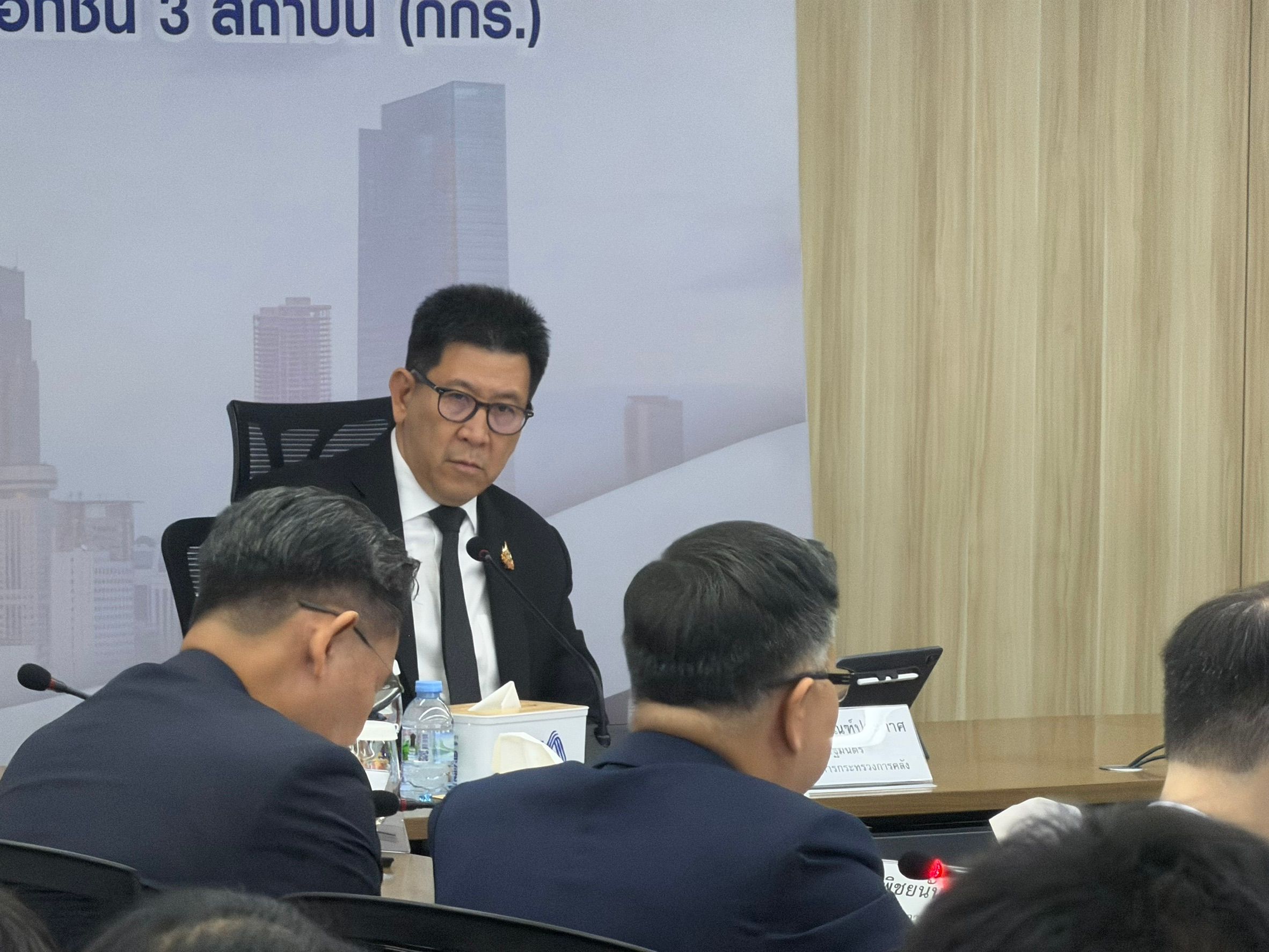A man in glasses and a suit sits at a meeting table, looking right.