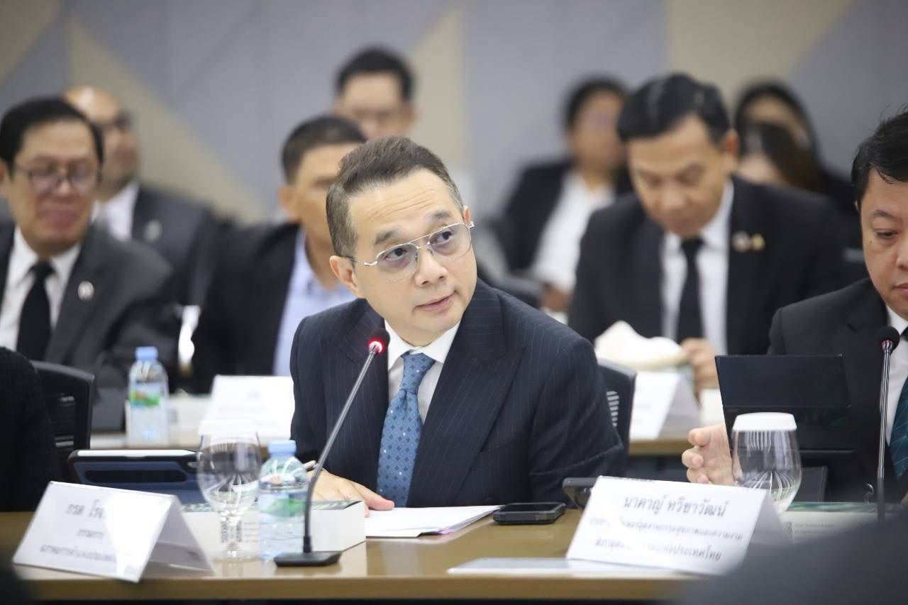 Man in dark suit and glasses sits at conference table with microphone.