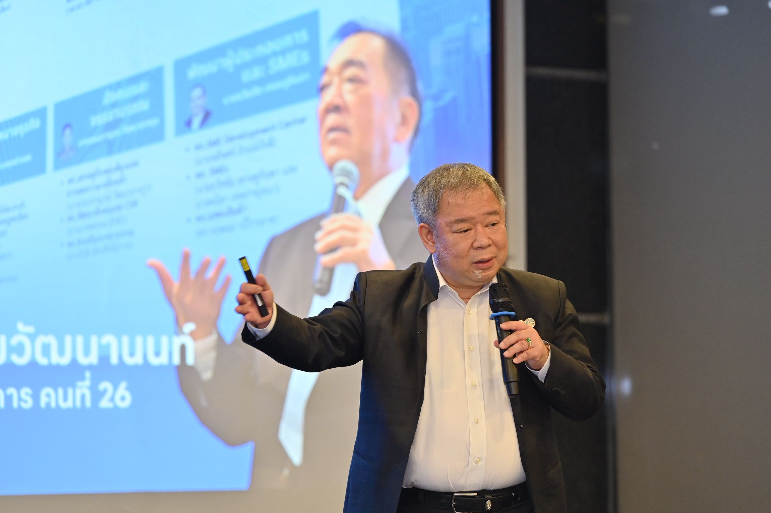 Man in suit speaking into microphone, gesturing towards a presentation screen.