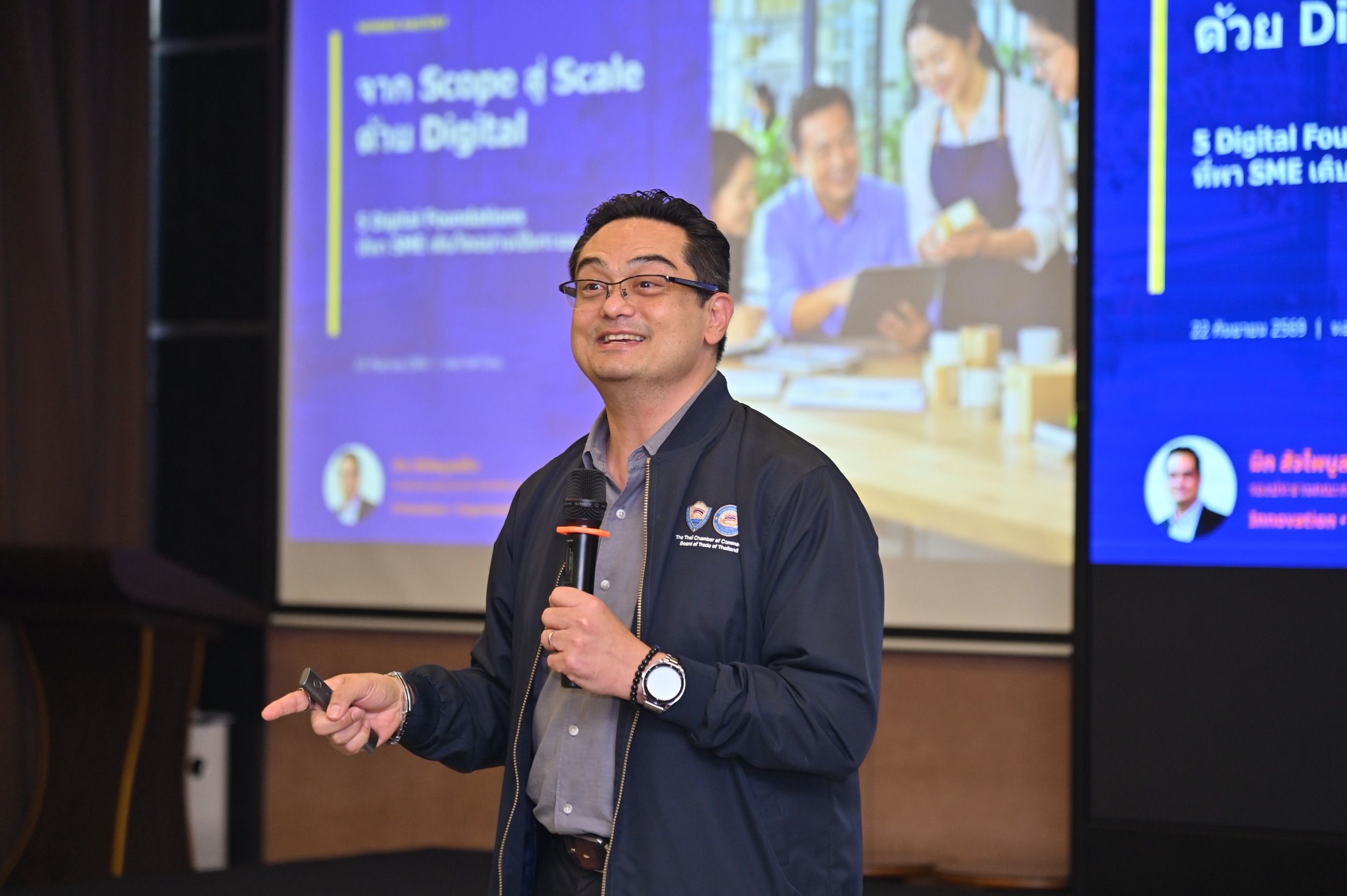 A man in glasses speaks into a microphone, presenting in front of a digital presentation screen.