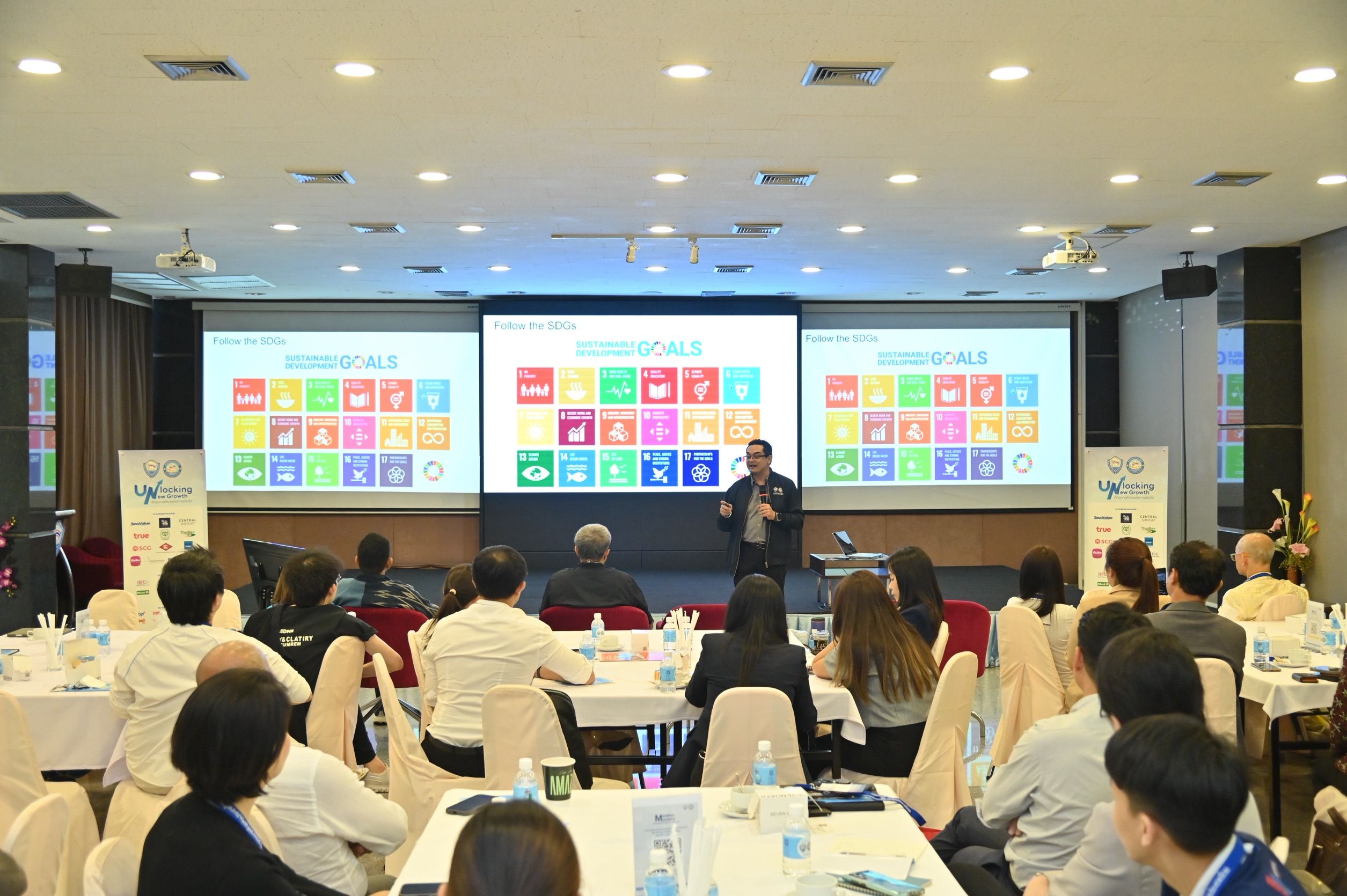 Presenter speaks to an audience in a conference room with screens displaying colorful charts.