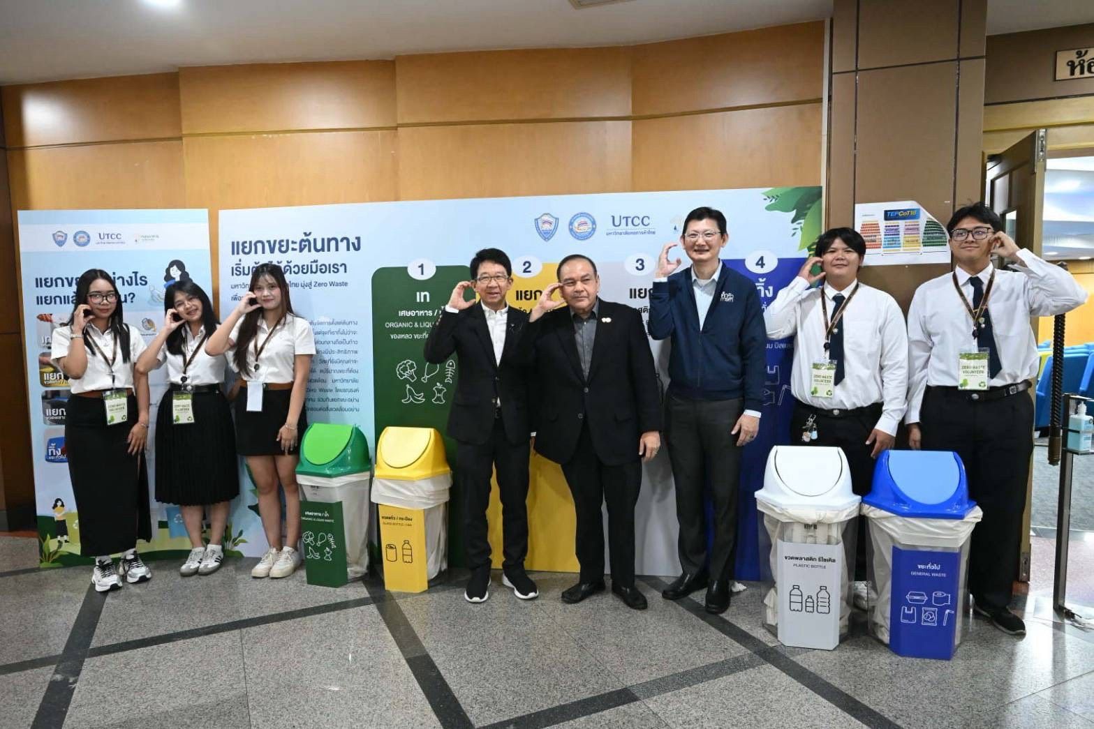 Students and adults stand with recycling bins, many saluting in front of a sustainability banner.