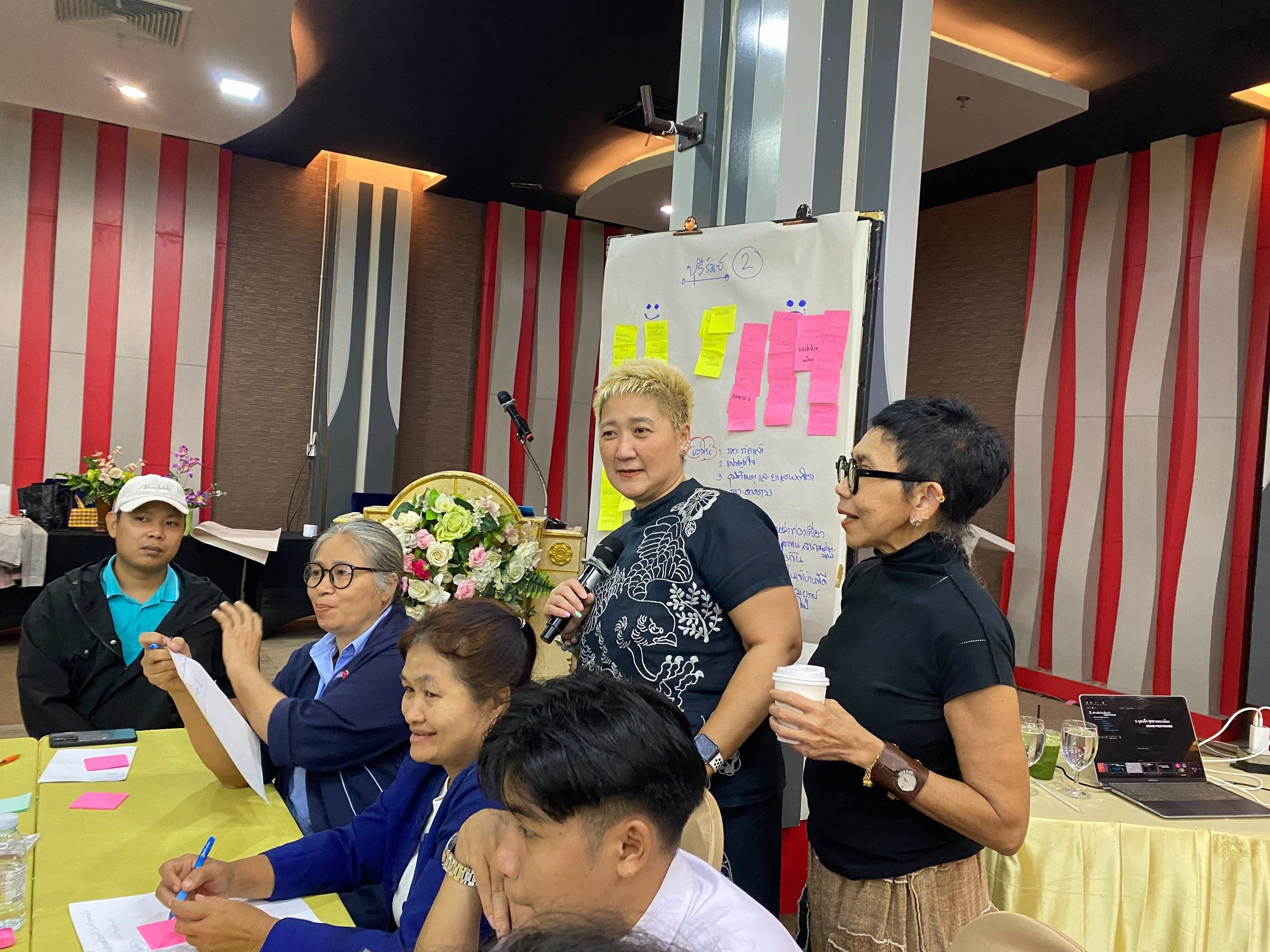 A woman presents to a diverse group in a meeting, with colorful notes visible.