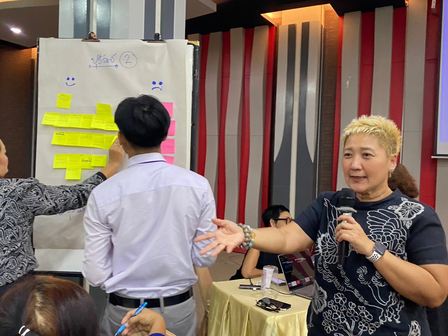 Woman speaking into microphone at a workshop with people interacting with a flip chart.
