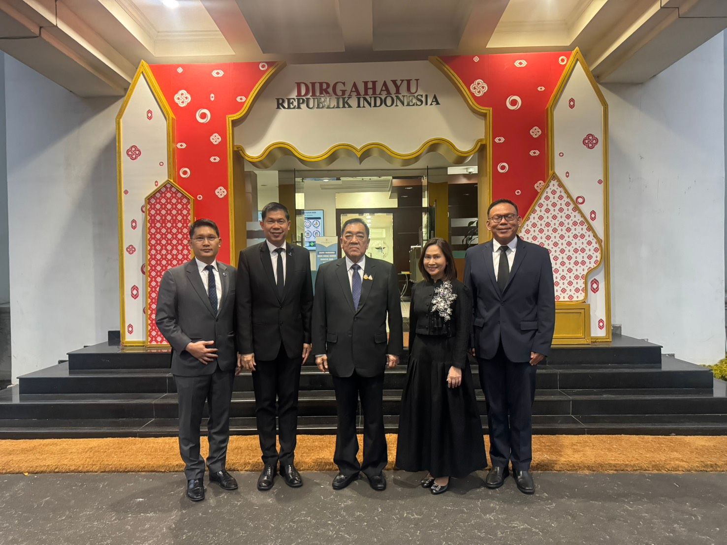 Five formally dressed individuals pose in front of a red and white archway celebrating Indonesia.