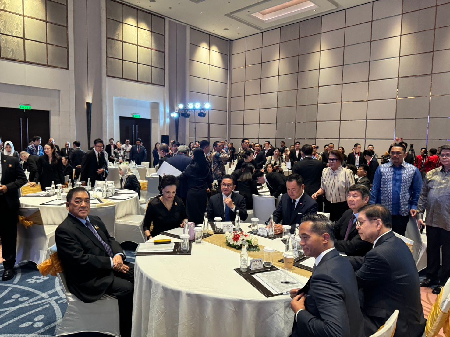 Attendees in business attire gather at round tables in a large conference hall.