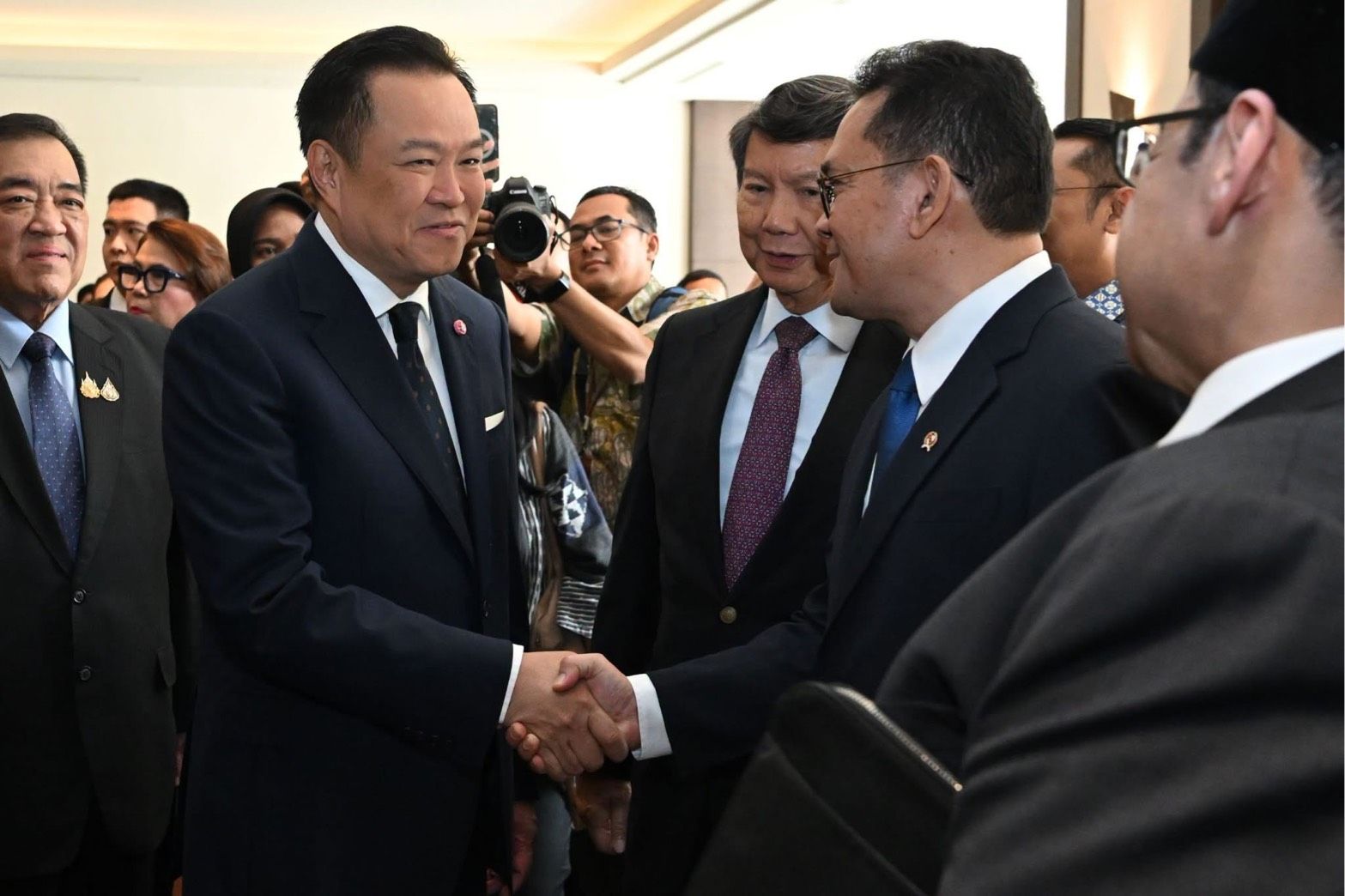 Two well-dressed men in dark suits shake hands at a formal indoor event.