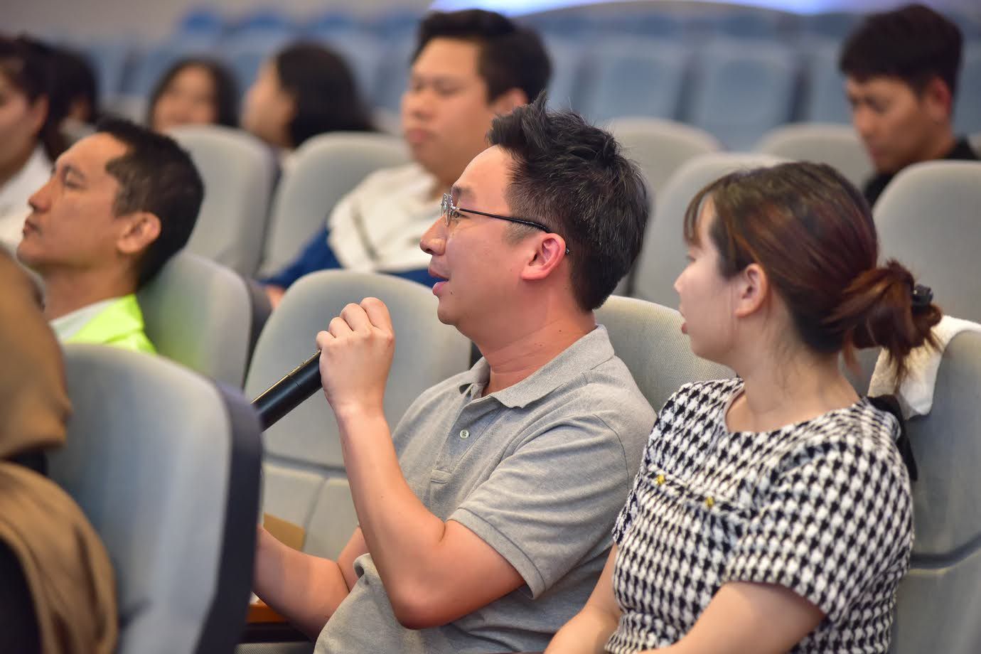 A man in glasses speaks into a microphone while seated in an audience.