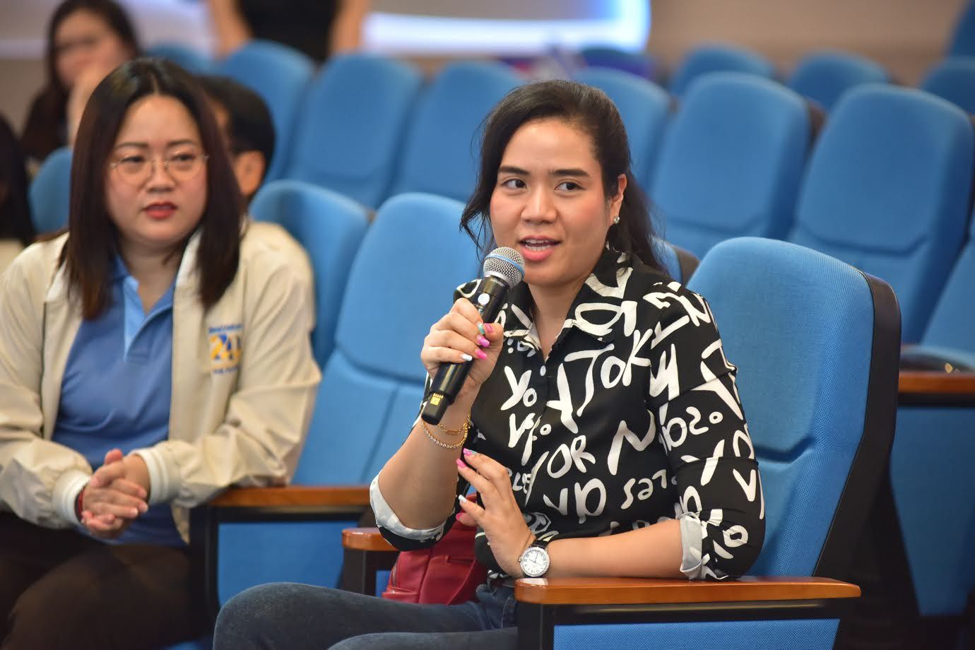 Woman in patterned shirt speaks into microphone during an auditorium event.
