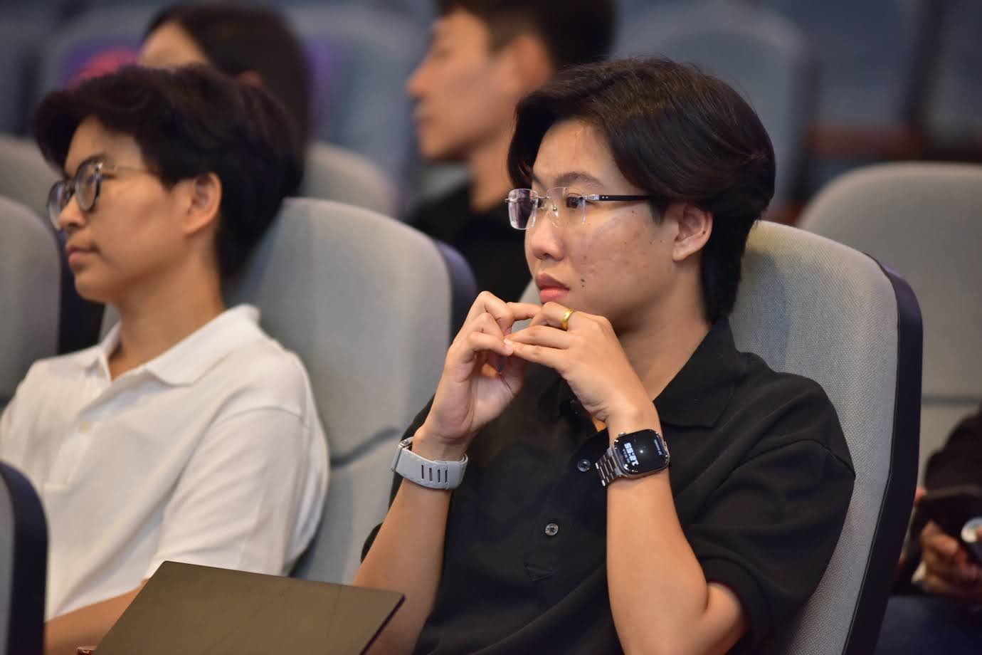 Young person in glasses and black shirt listening intently in a lecture hall.